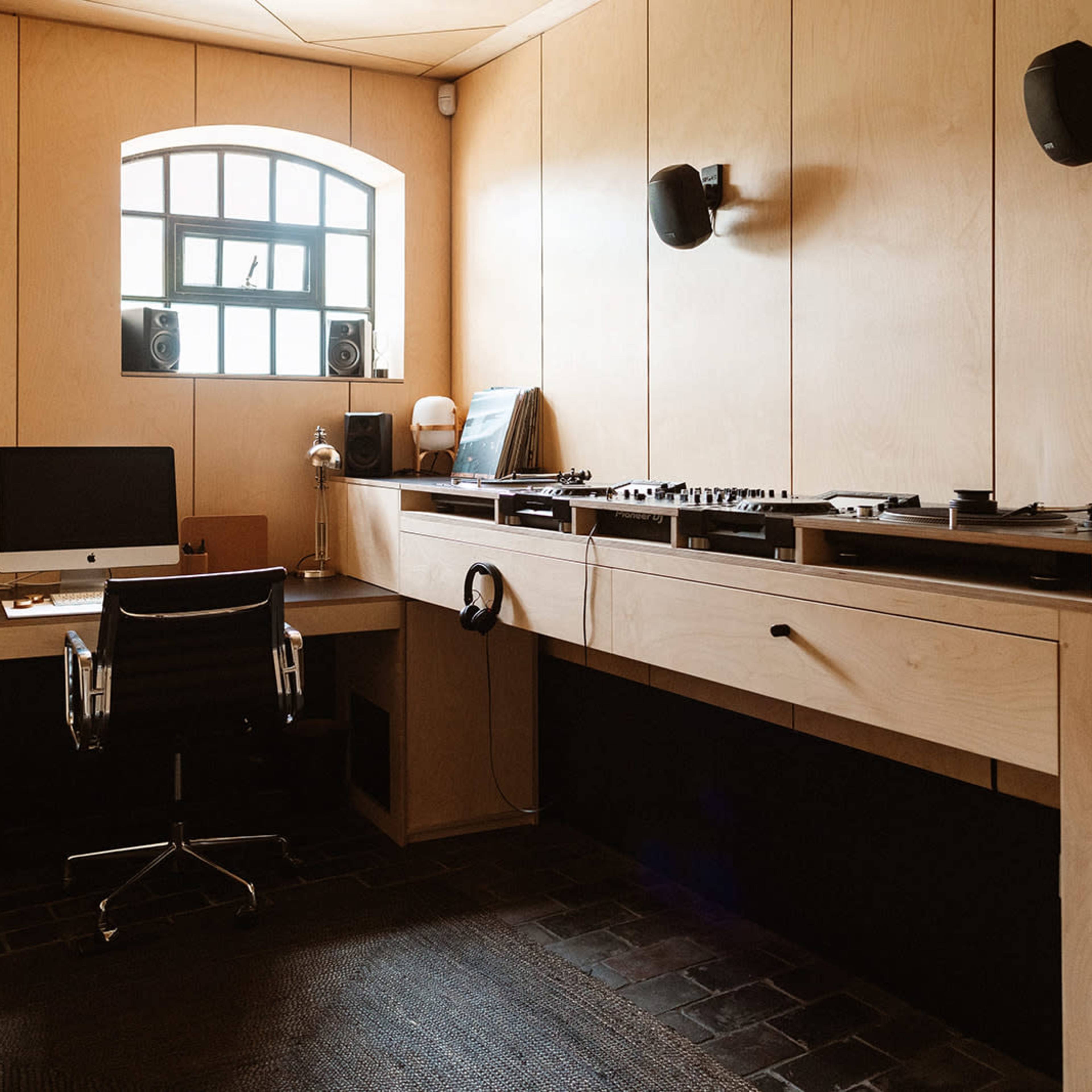 A minimalist office space features a large wooden desk with a computer, audio equipment, and a wall-mounted shelf beside a window.