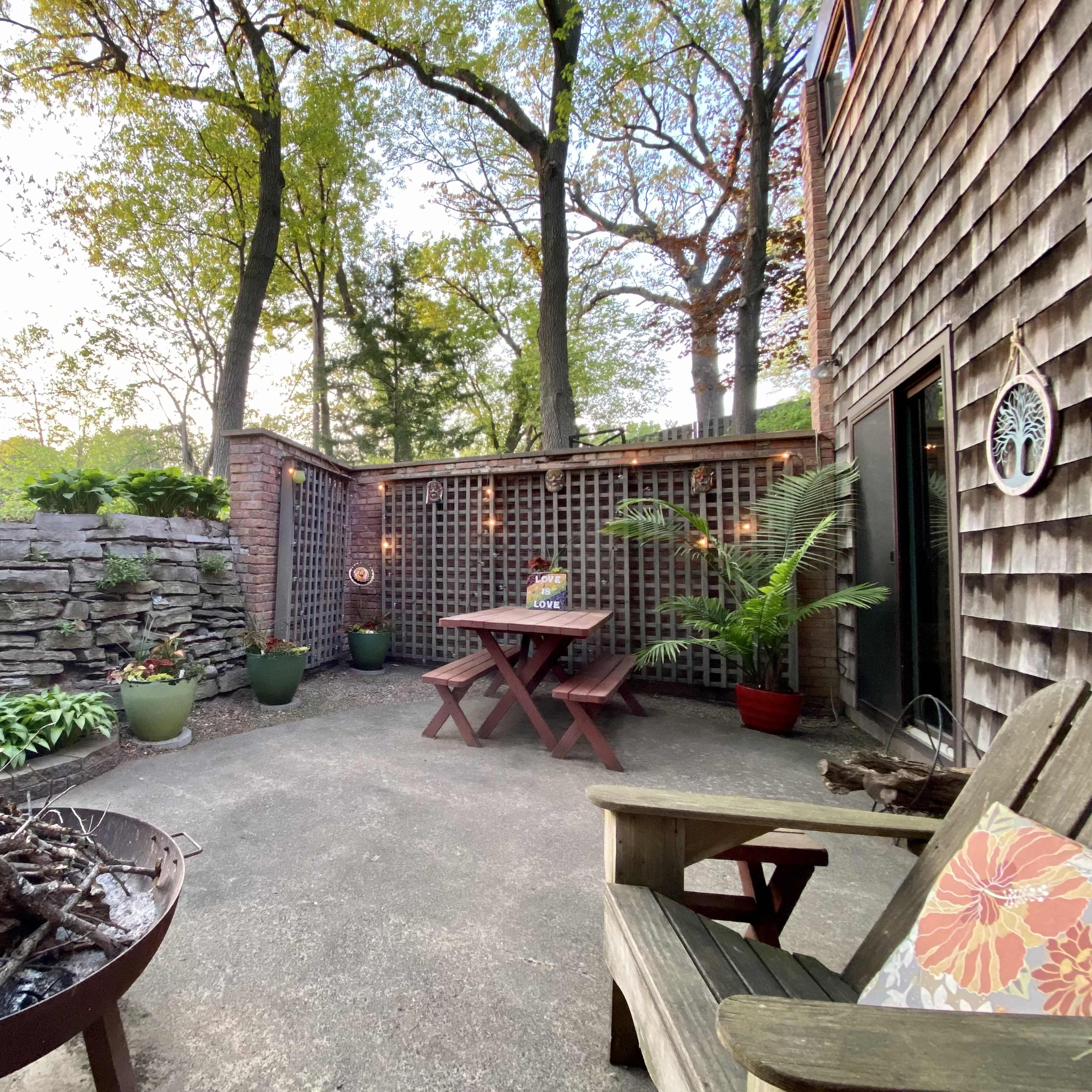 A patio with a fire pit surrounded by plants, a wooden table, and a chair, all set against a backdrop of trees and a stone wall.