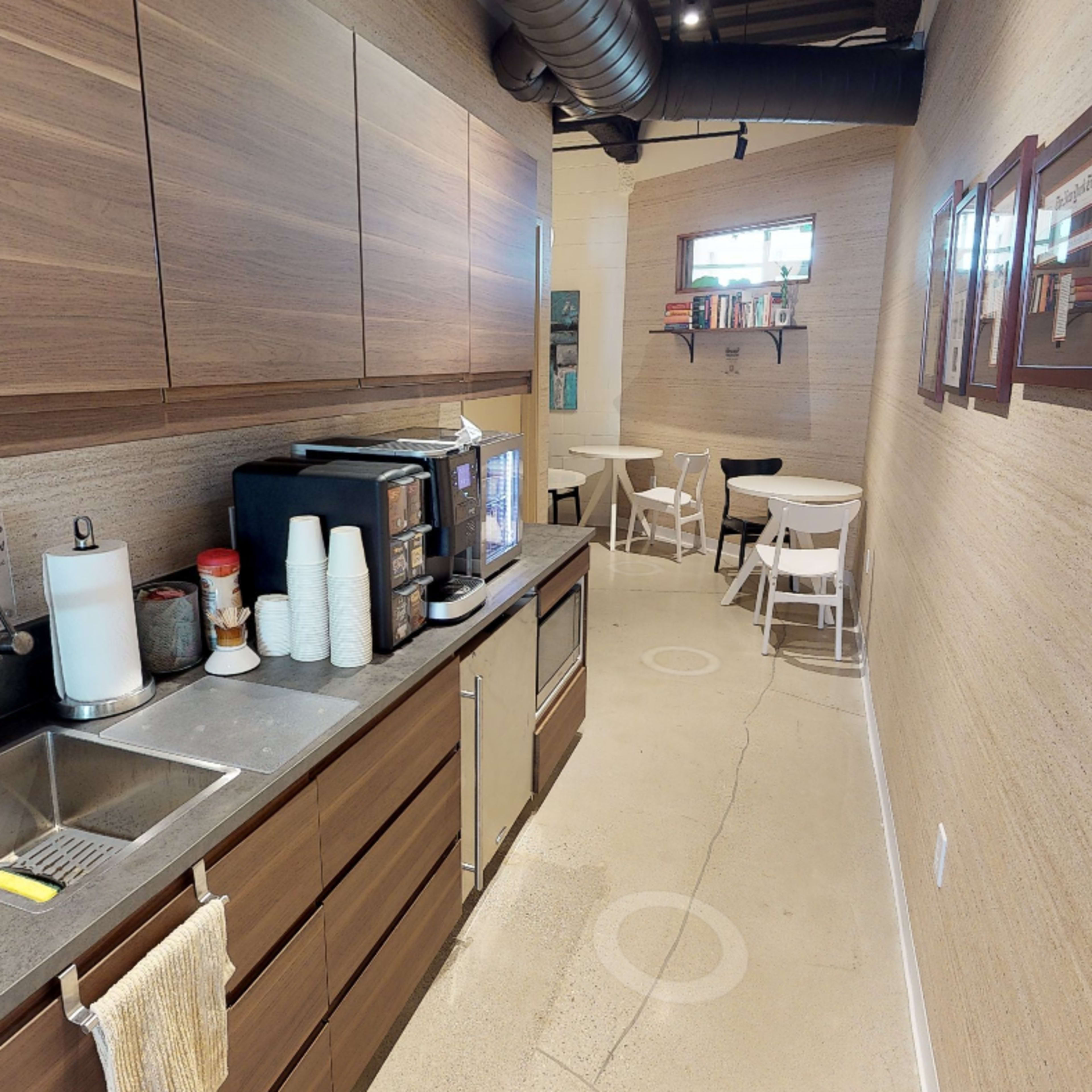 A narrow kitchen area with wooden cabinets, a sink, and a coffee machine on one side, and a small seating area with white tables and chairs on the other.