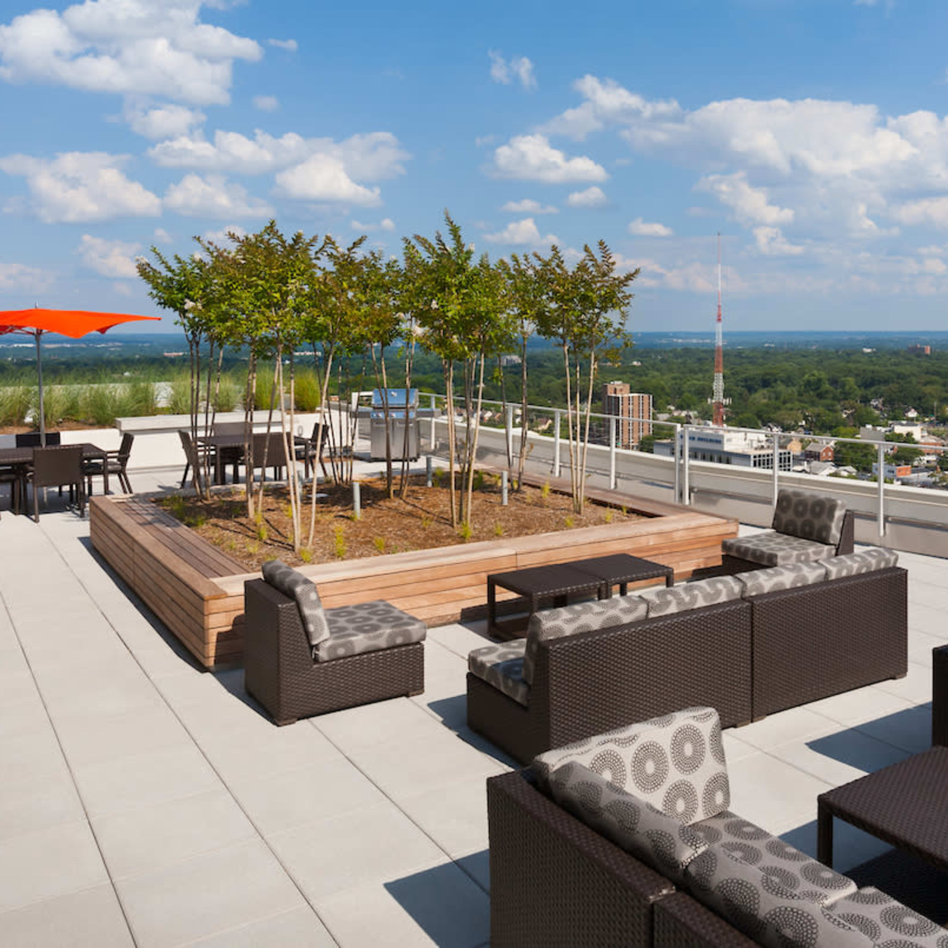 A rooftop terrace featuring modern furniture, a wooden planter with trees, and an orange umbrella, overlooking a cityscape under a partly cloudy sky.