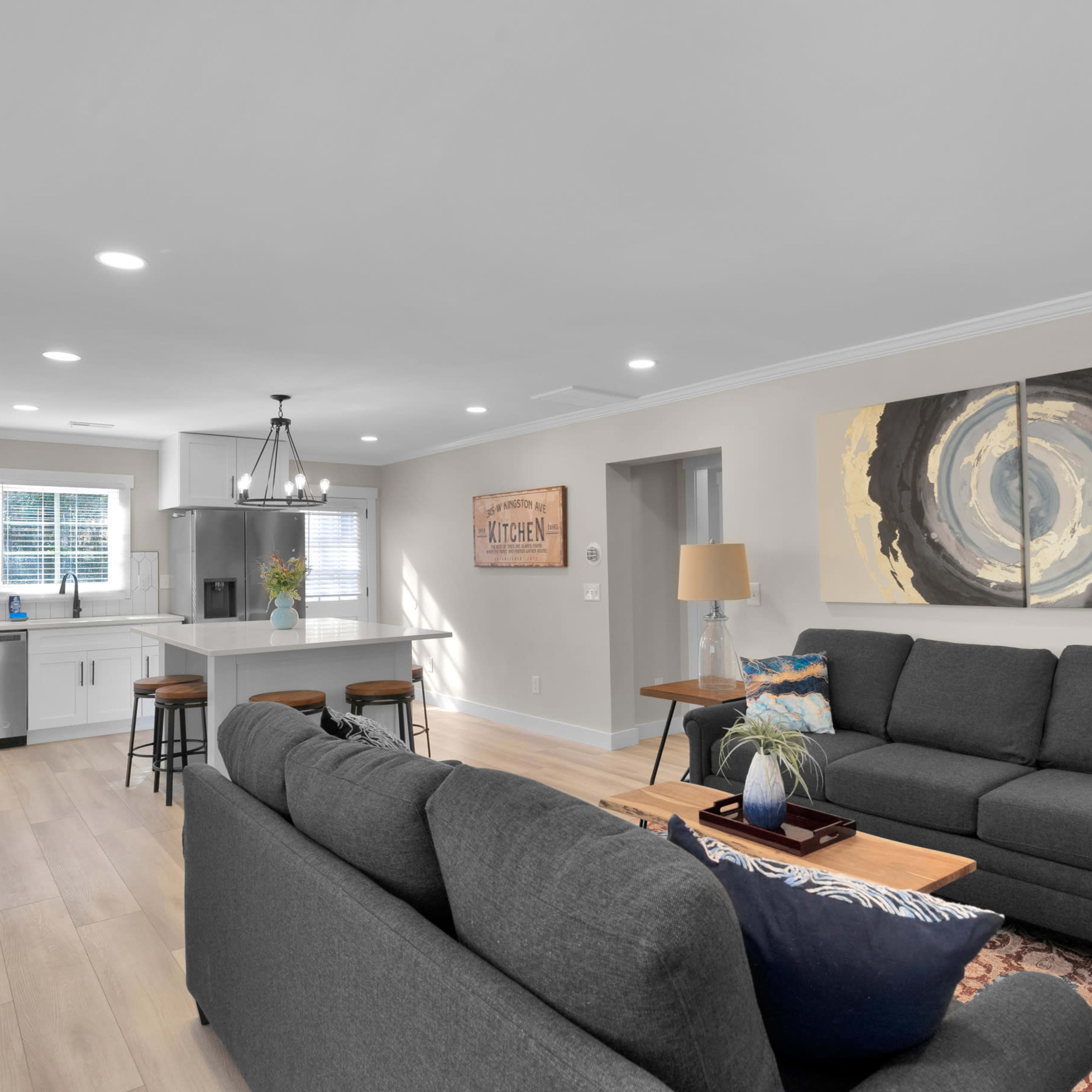 A modern living room with gray sofas, a wooden coffee table, and a kitchen area in the background featuring stainless steel appliances.