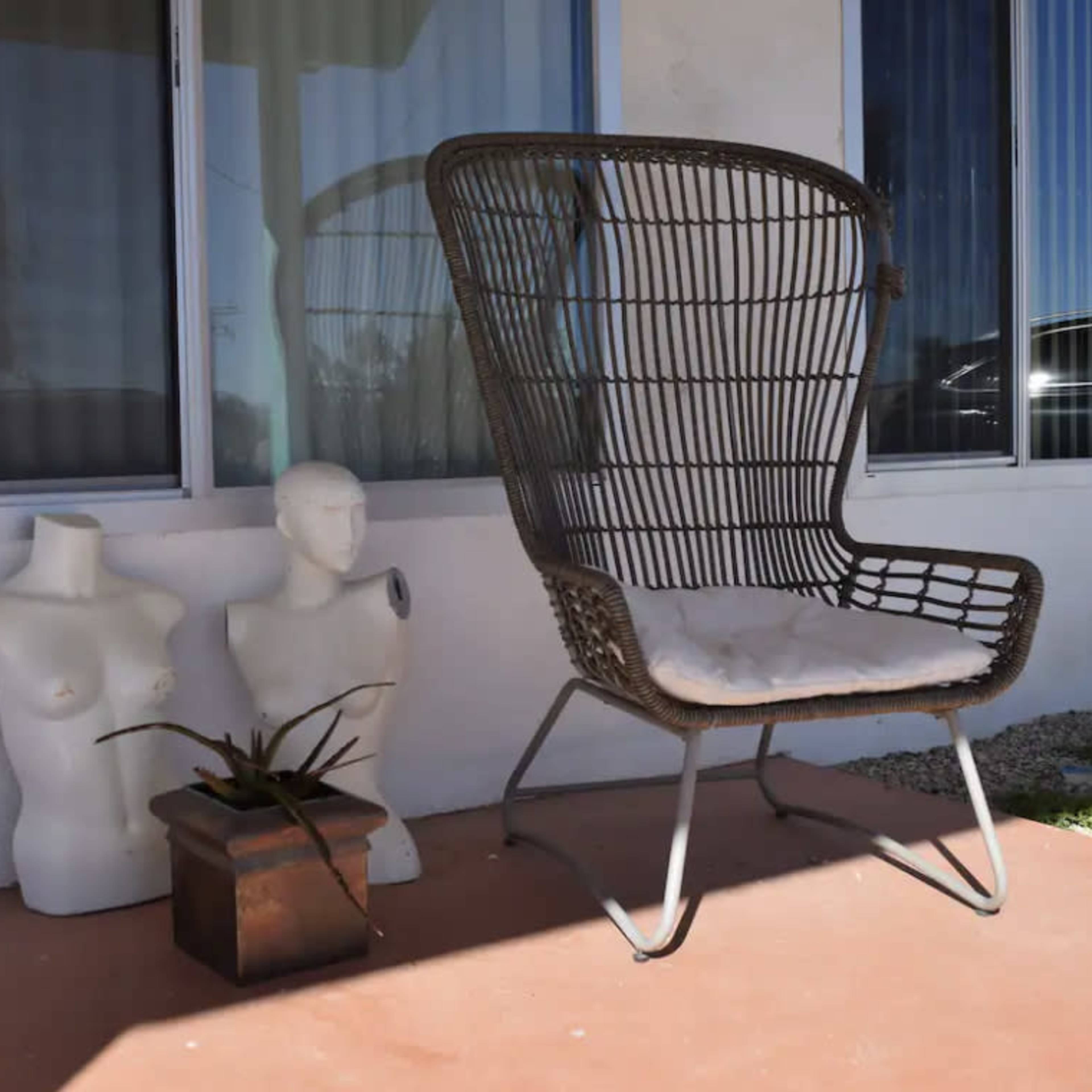 A large wire chair sits on a porch next to two mannequin torsos and a potted plant.