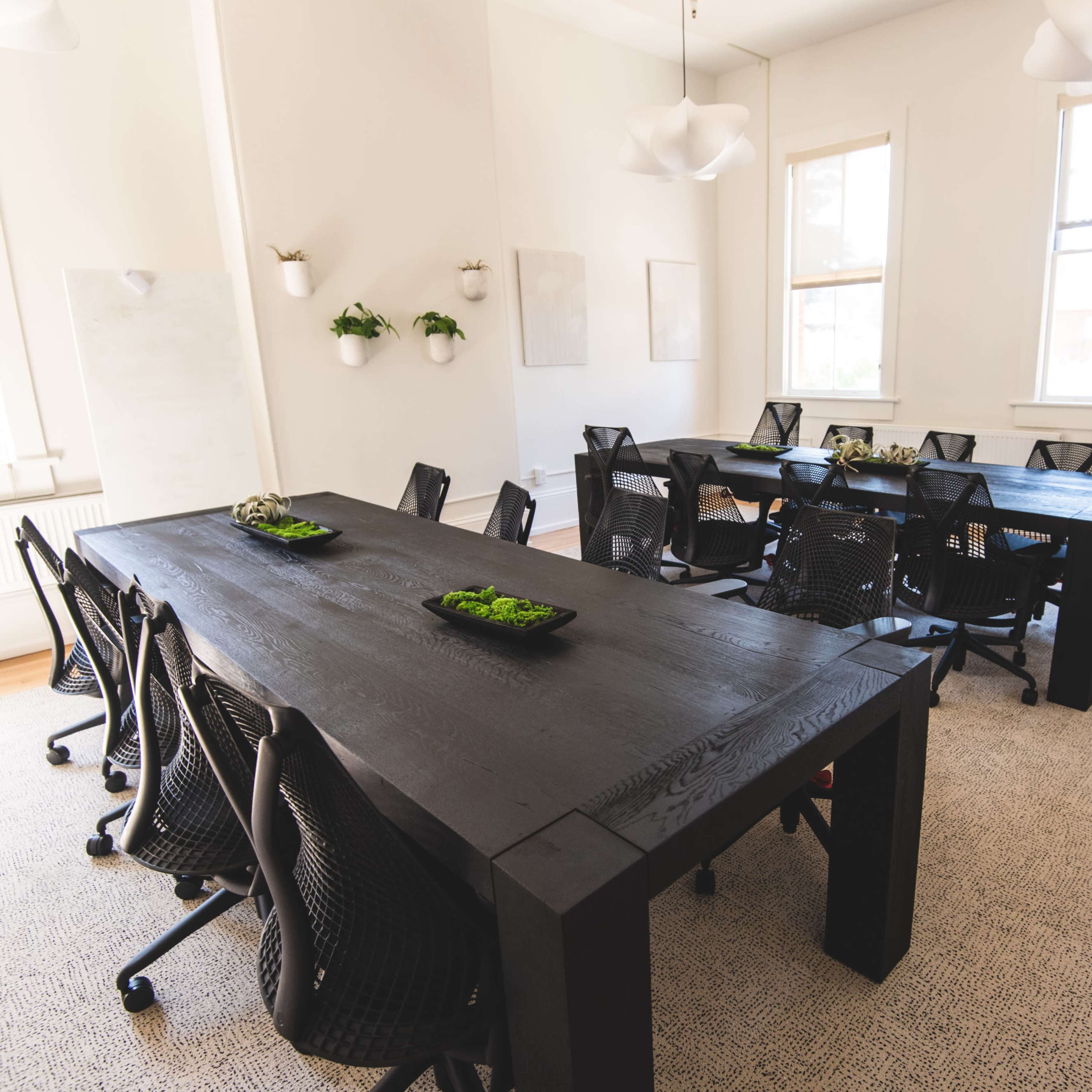 A bright meeting room features two large black tables surrounded by mesh office chairs and potted plants on the walls.