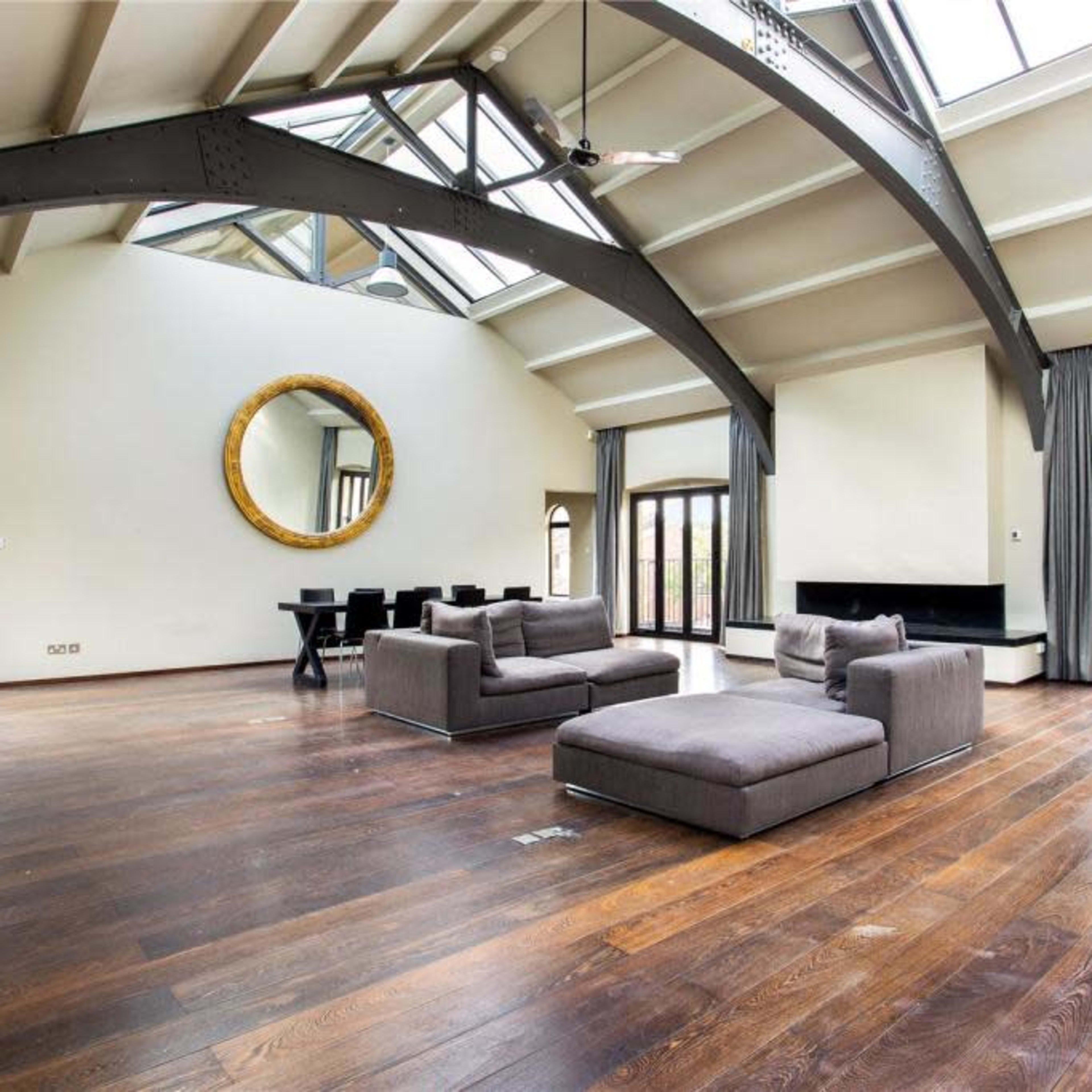 A spacious living room features a high ceiling with skylights, two gray sofas, a round mirror, and large glass doors leading to an outdoor area.