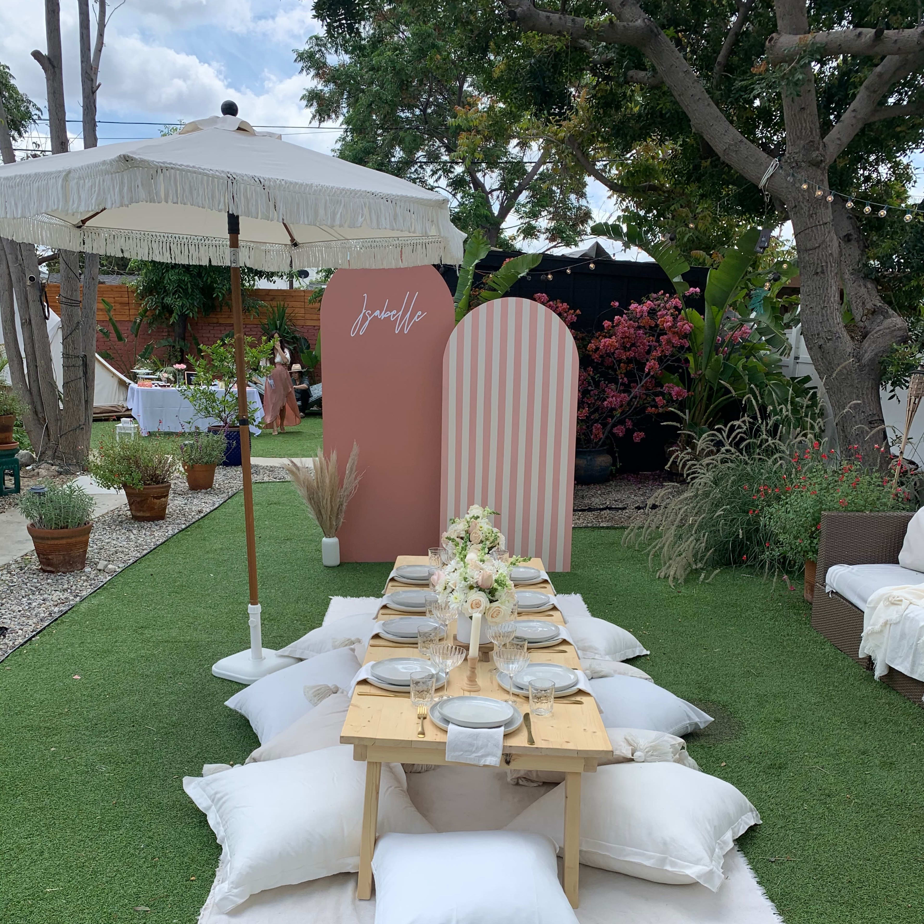 A low table set with plates and cutlery is placed on a white rug surrounded by pillows, with a decorative backdrop and an umbrella in an outdoor garden setting.