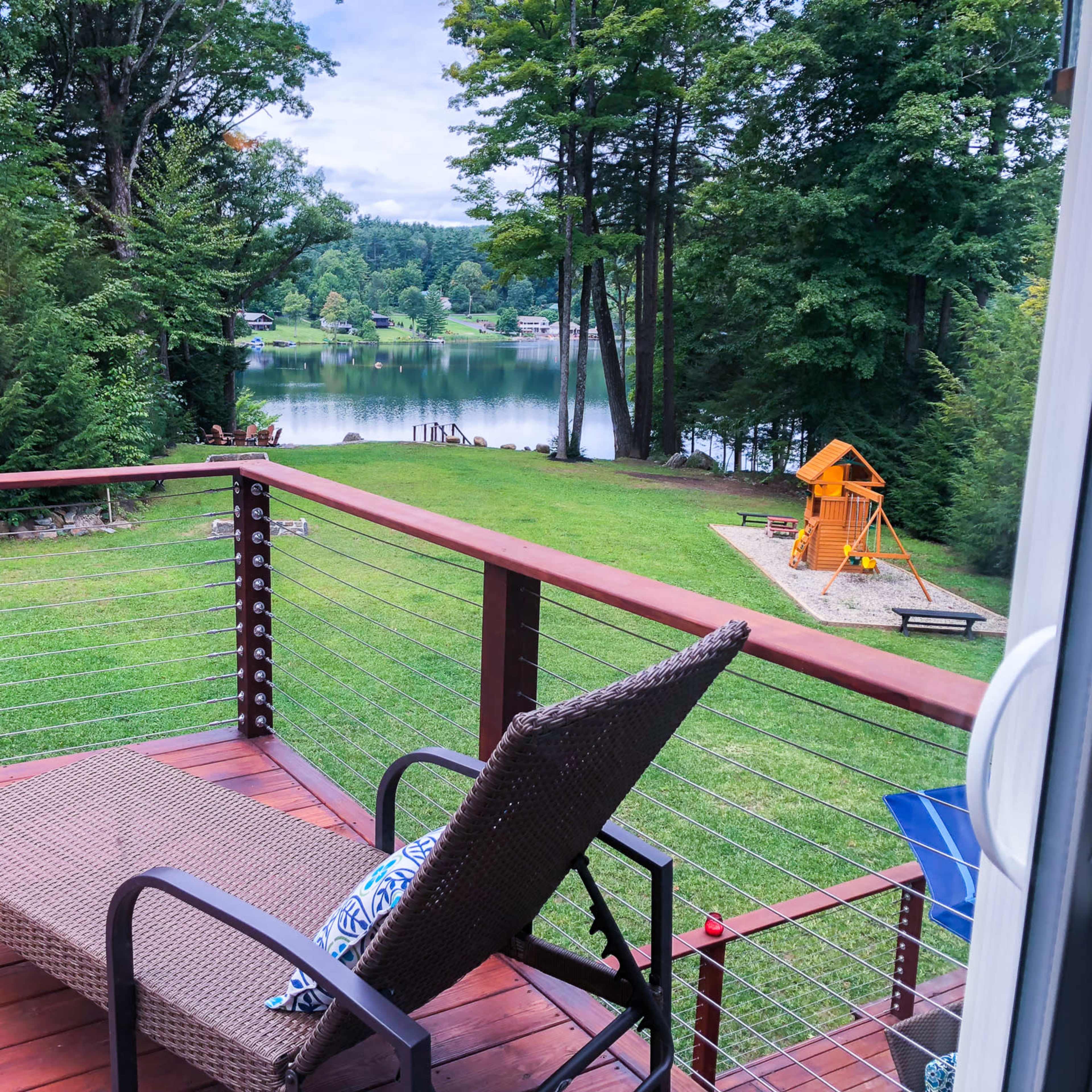 A wooden deck overlooks a tranquil lake surrounded by trees, with a playground visible in the background.
