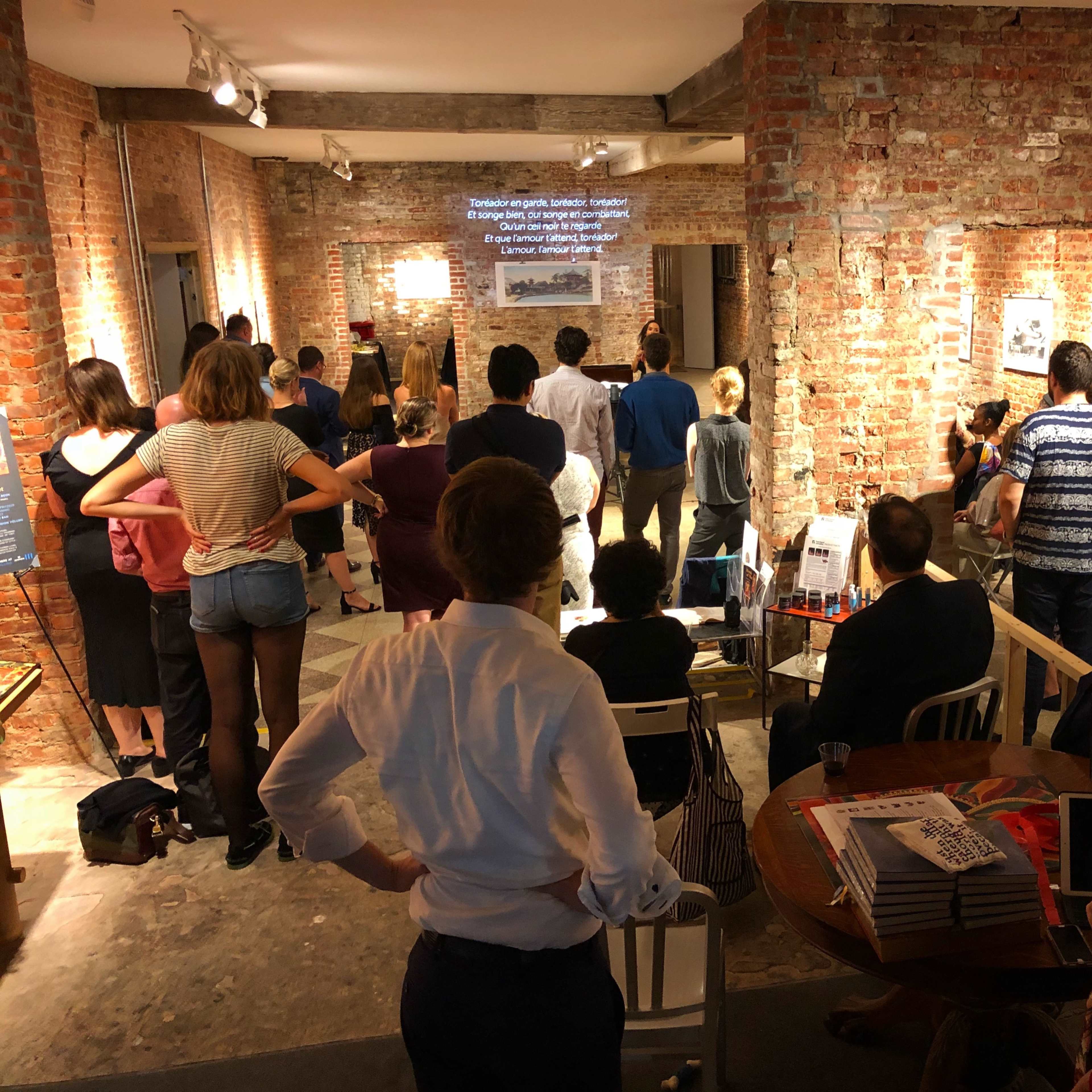 A group of people is gathered in an art space with exposed brick walls, watching a presentation on a screen.