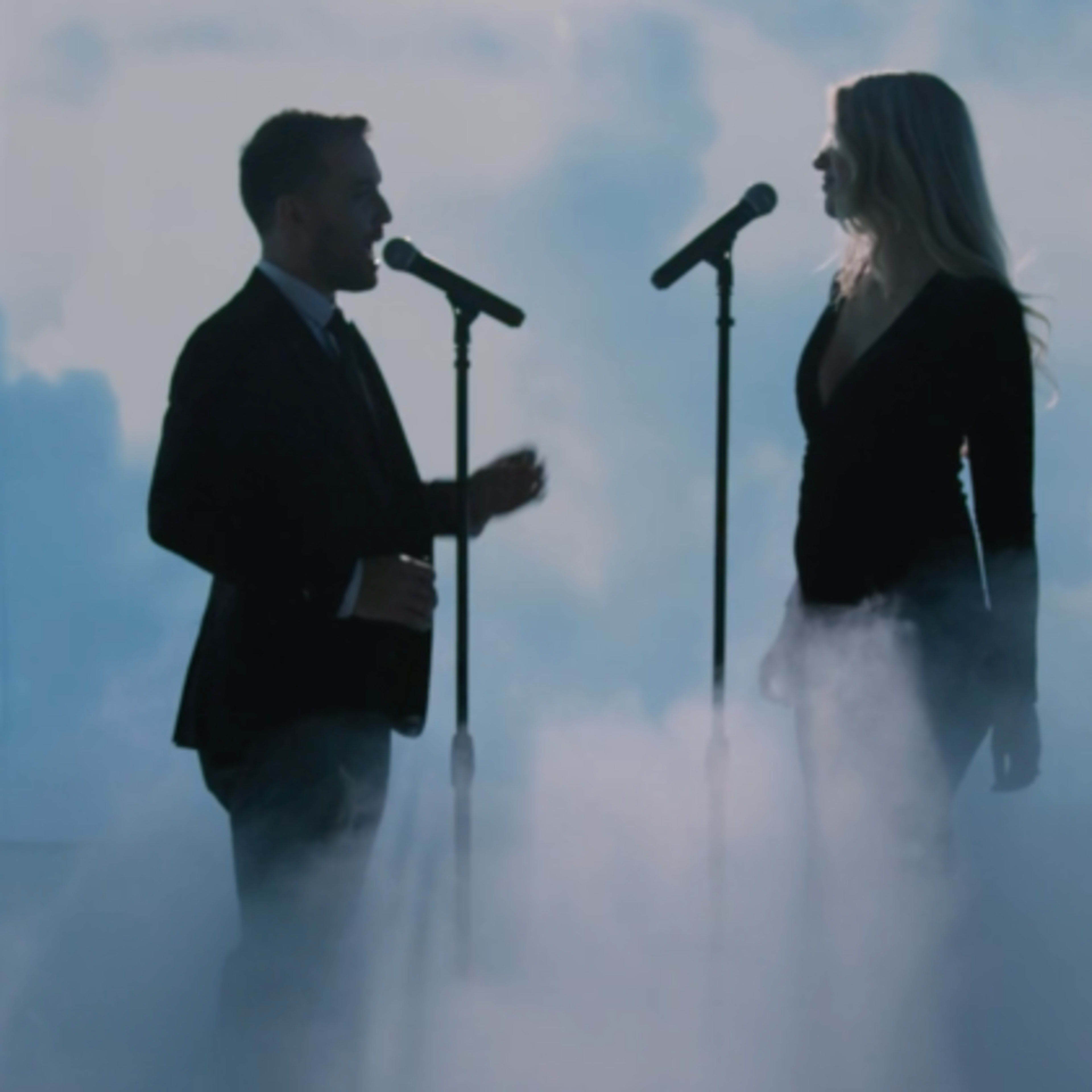 Two singers perform on stage in front of microphones, surrounded by a backdrop of clouds and fog.