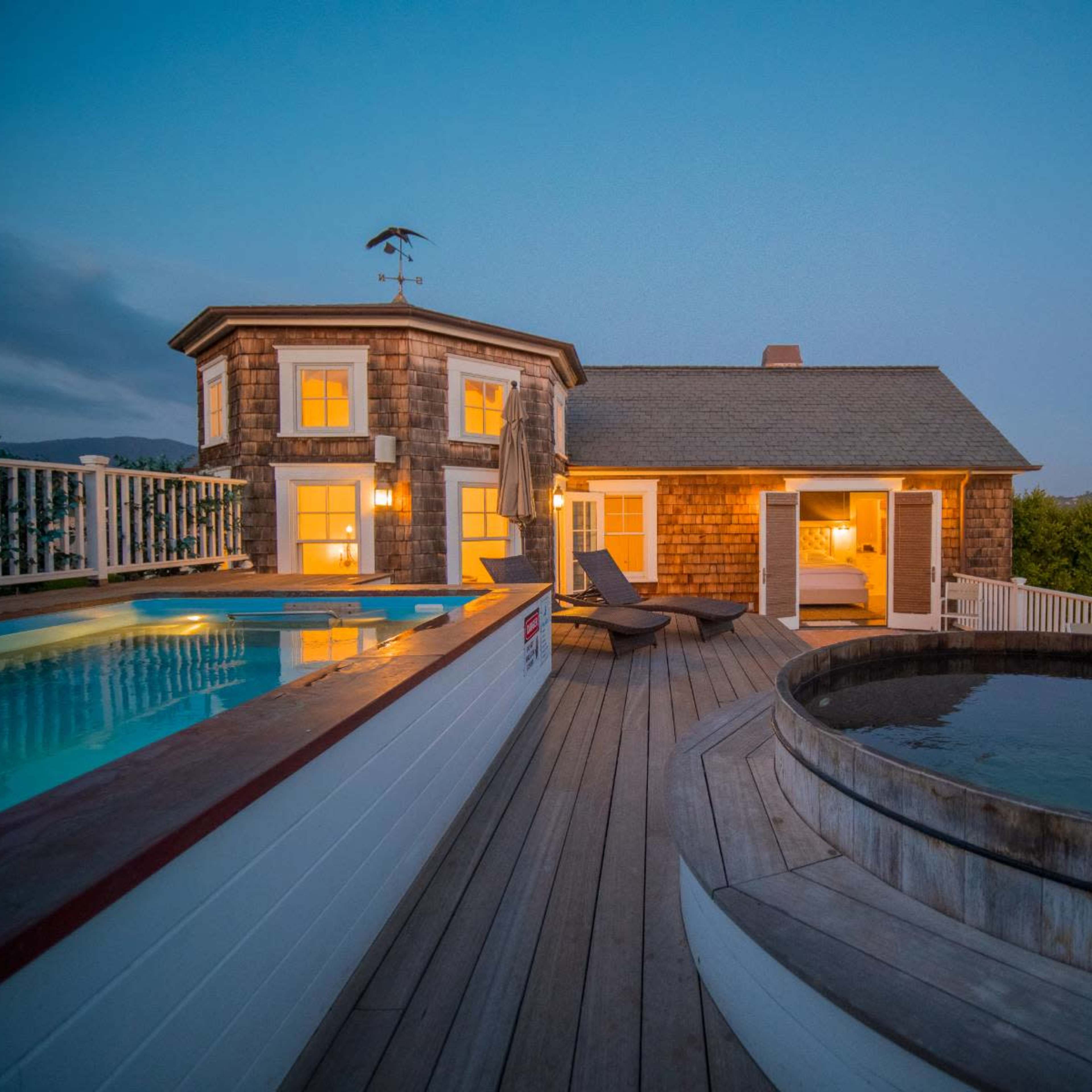 A waterfront property features a hot tub and swimming pool on a wooden deck overlooking the water at dusk.