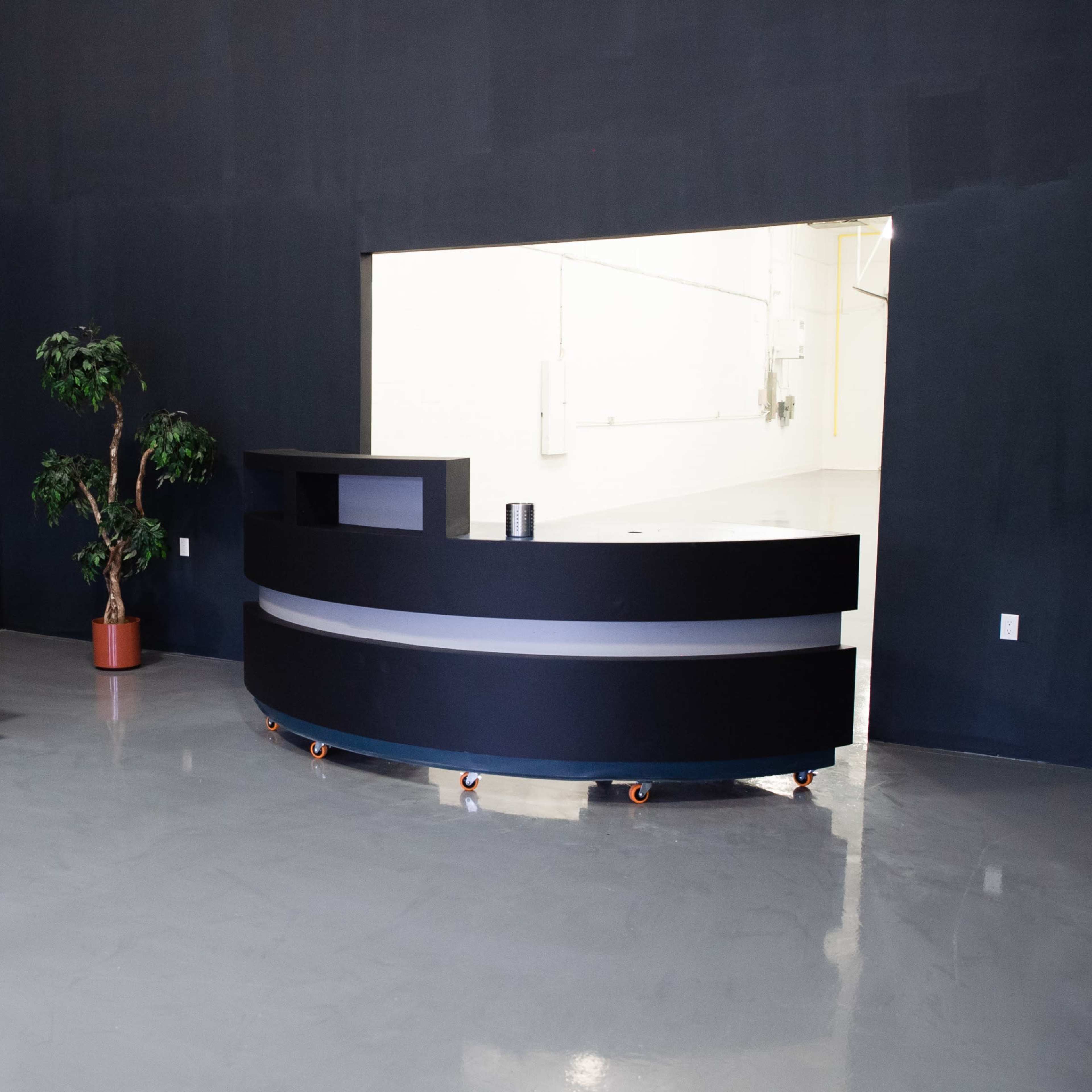 An empty room with a black wall featuring a reception desk, a wooden table and chairs, and a potted plant near the entrance.