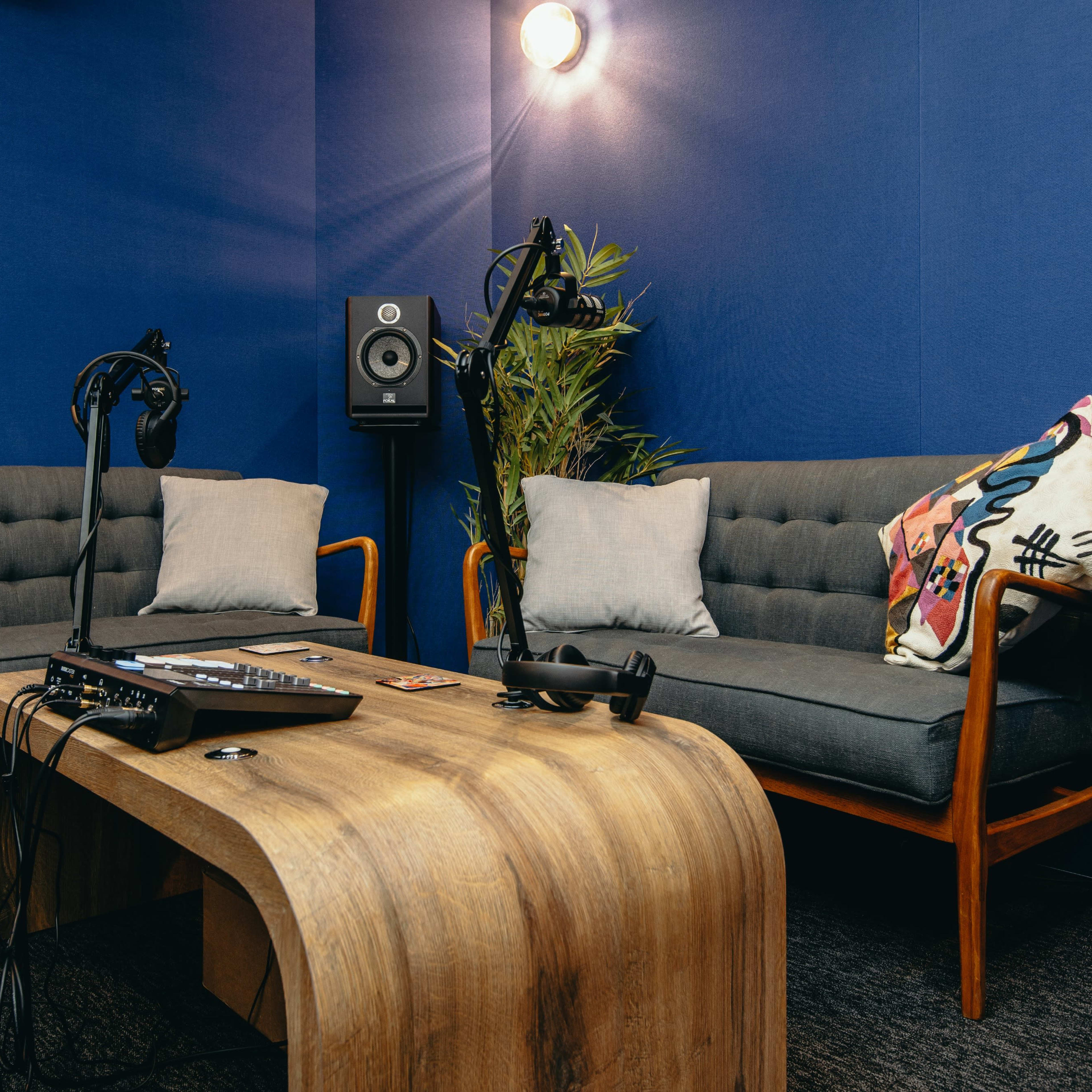 A small recording studio with two gray couches, a wooden table, microphones, and audio equipment against a blue wall.