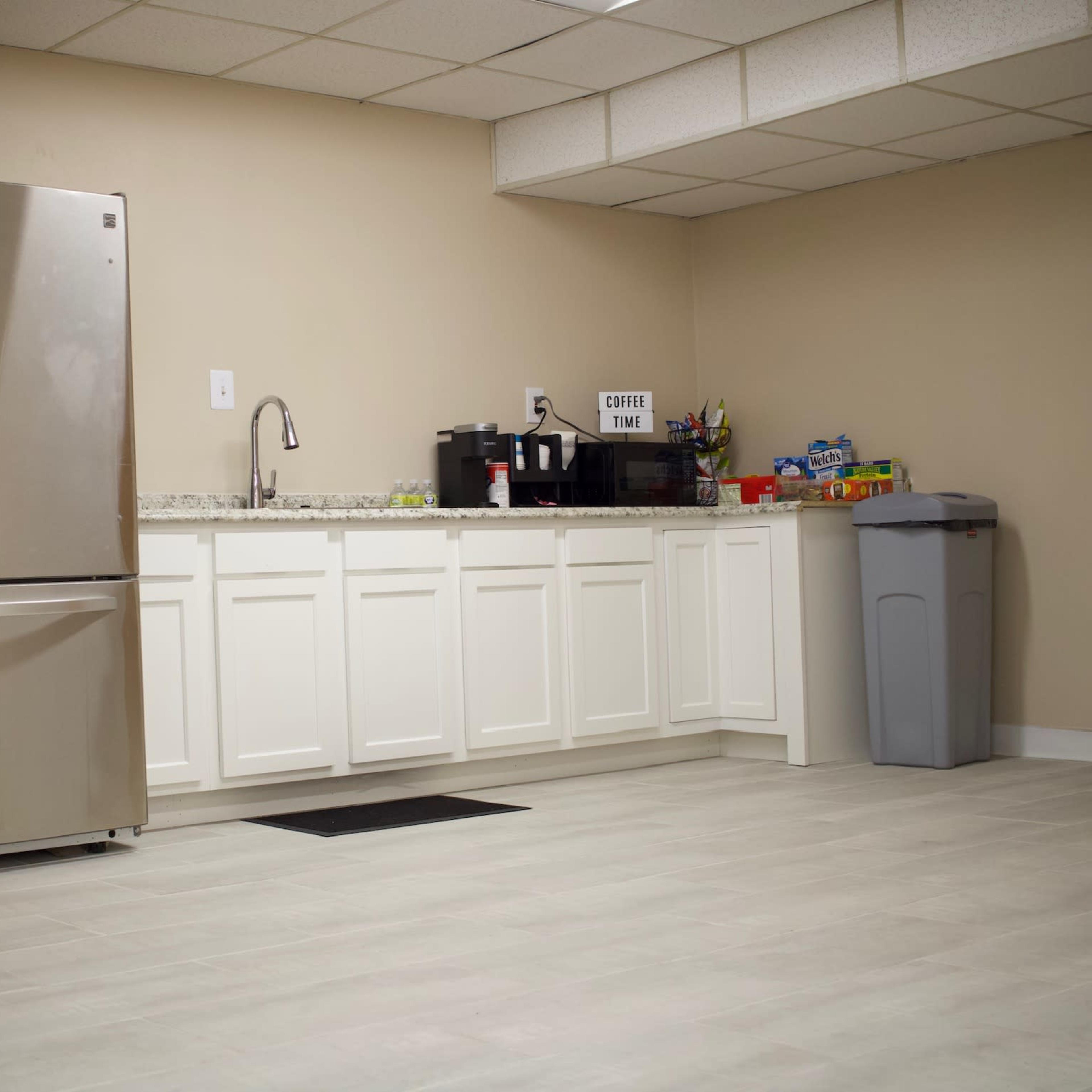 A stainless steel refrigerator and coffee station are placed against a beige wall in a simple kitchen with tiled flooring.