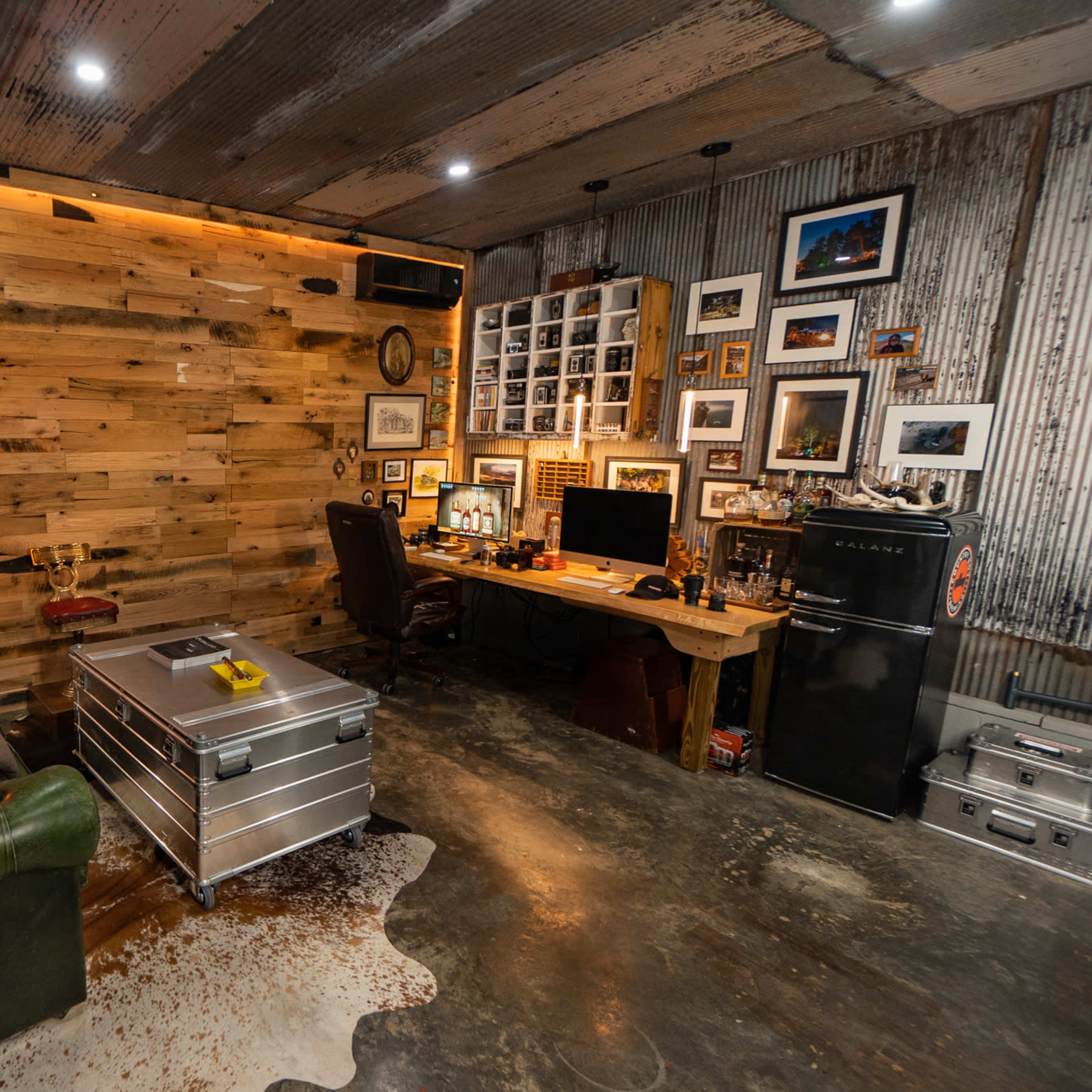 A rustic office space featuring a wooden accent wall, a desk with a computer, a vintage-style fridge, and a green leather couch.