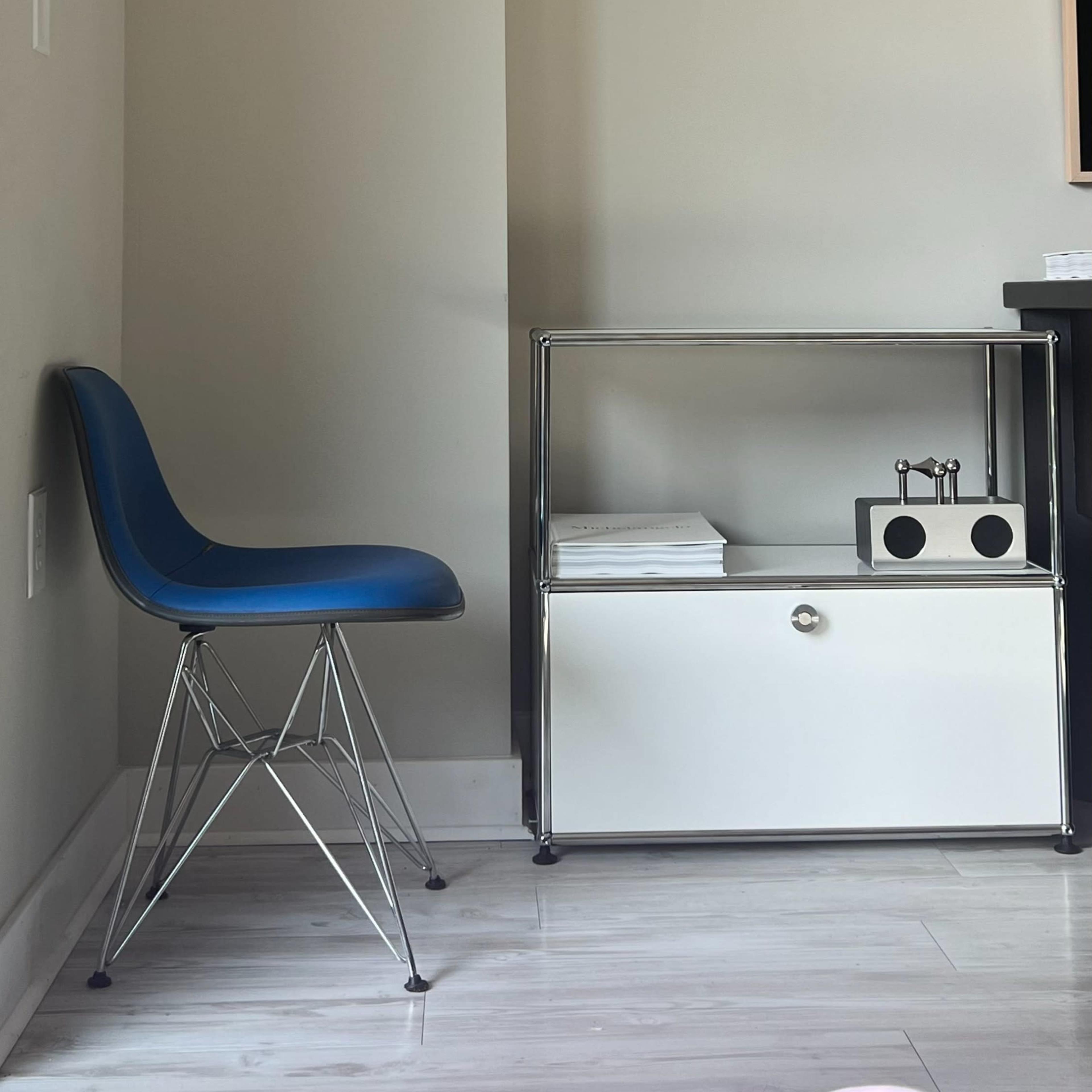A blue chair is placed next to a white shelf with a black and white decorative piece against a light gray wall, featuring a portrait hanging above.
