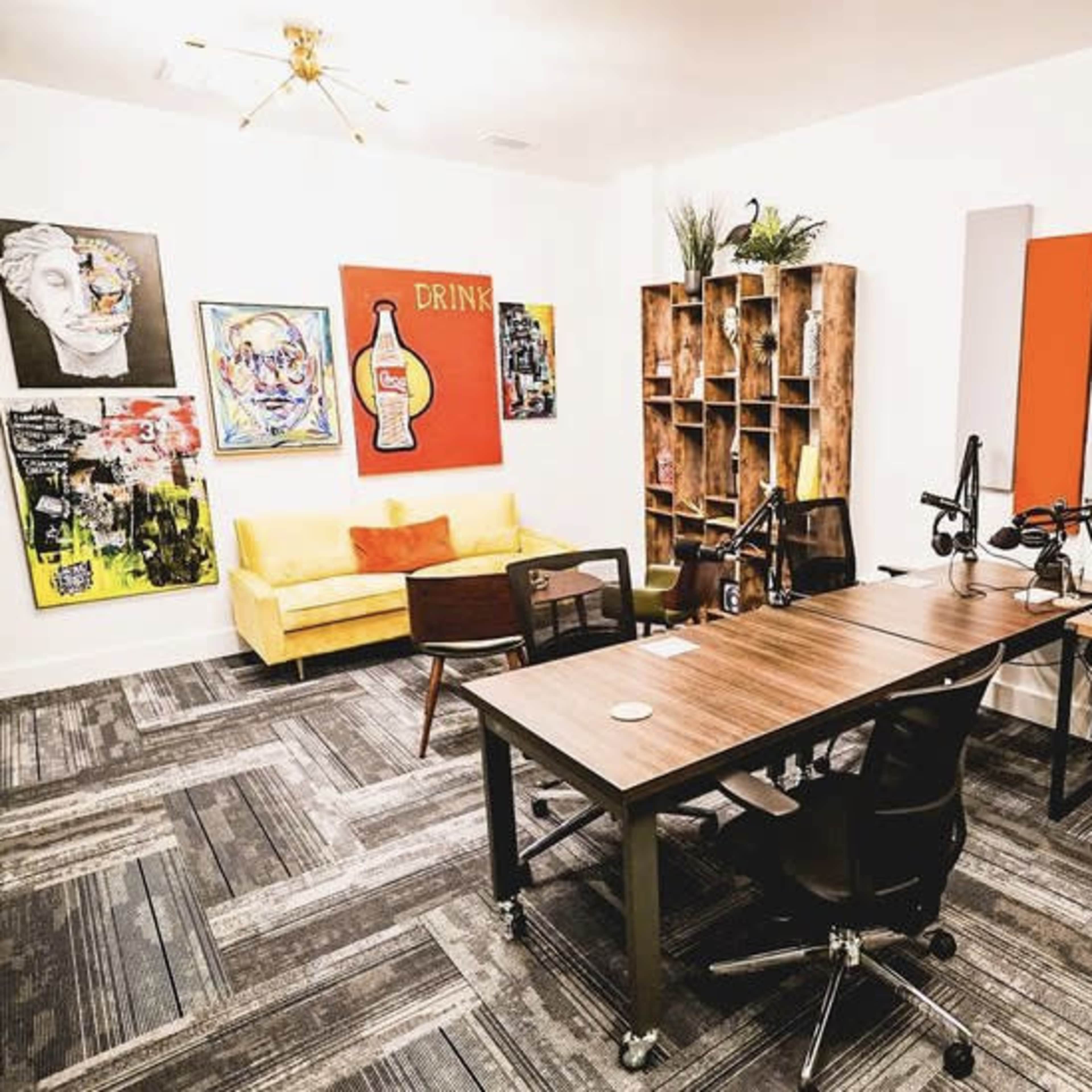 A modern workspace featuring a sofa, artwork on the walls, a wooden bookshelf, and desks equipped with microphones and computers.