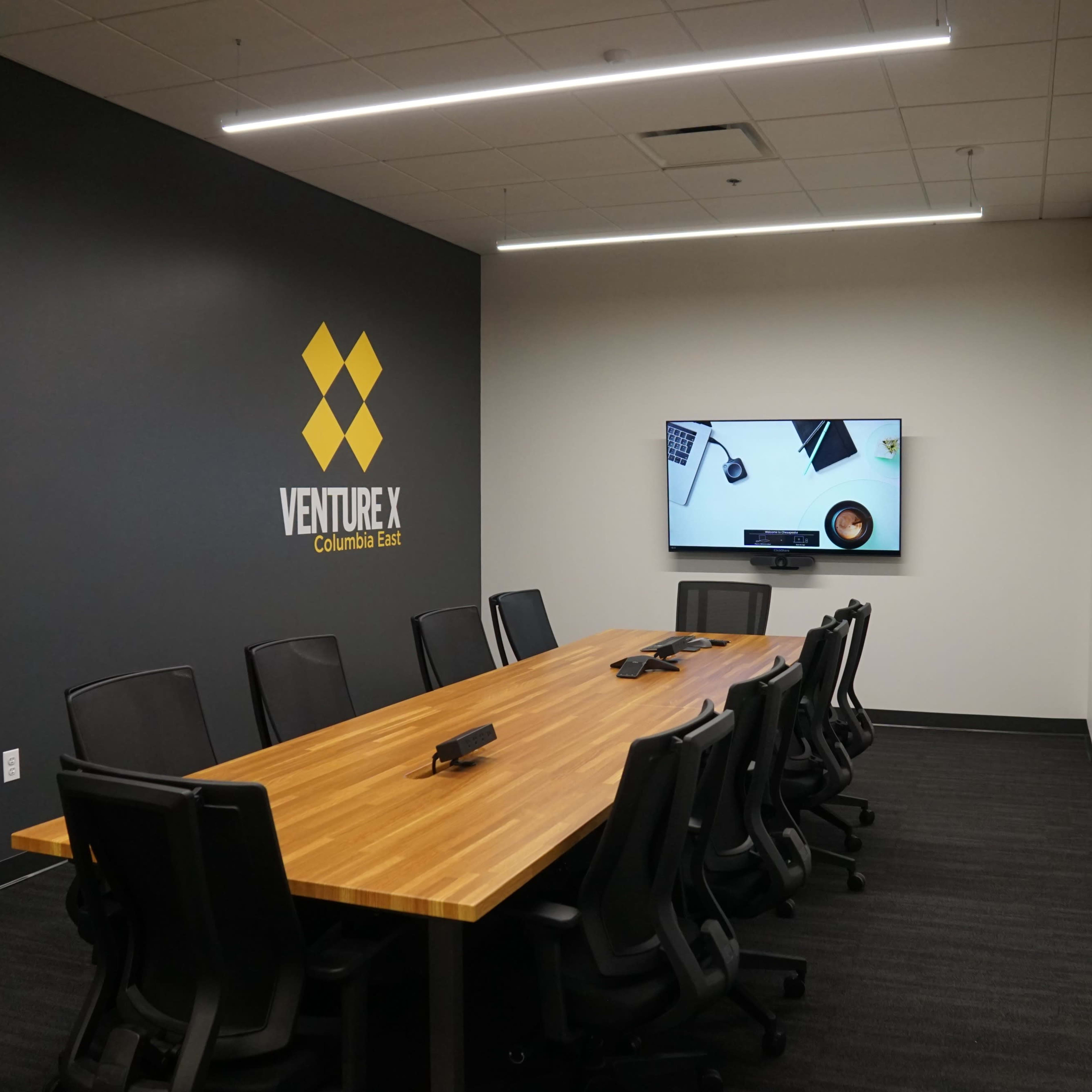 A modern conference room with a long wooden table, black ergonomic chairs, a large wall-mounted TV, and the "Venture X" logo displayed on the dark wall.