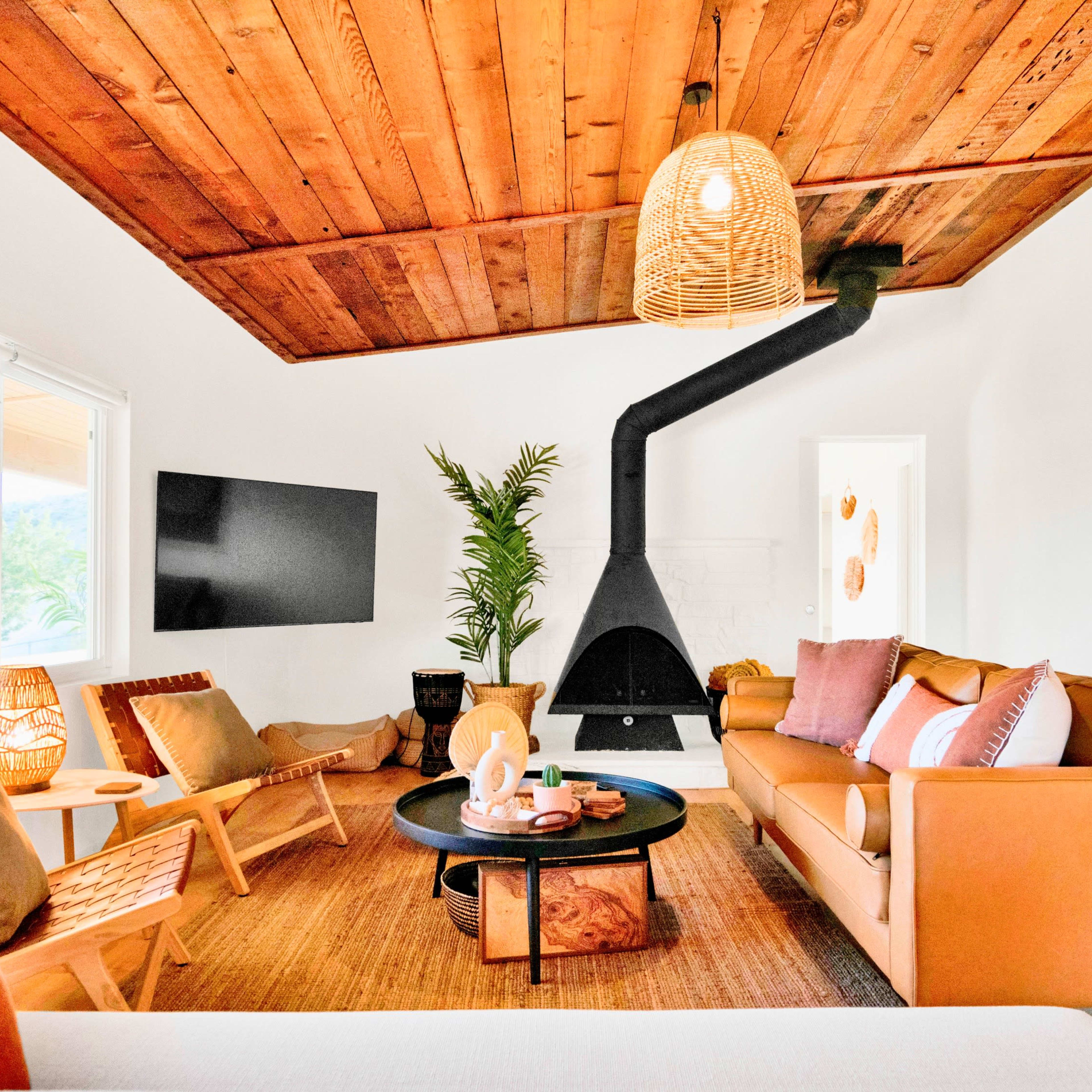A modern living room features a leather sofa, mid-century chairs, a round coffee table, and a prominent black fireplace, all under a wooden ceiling and accompanied by tropical plants.