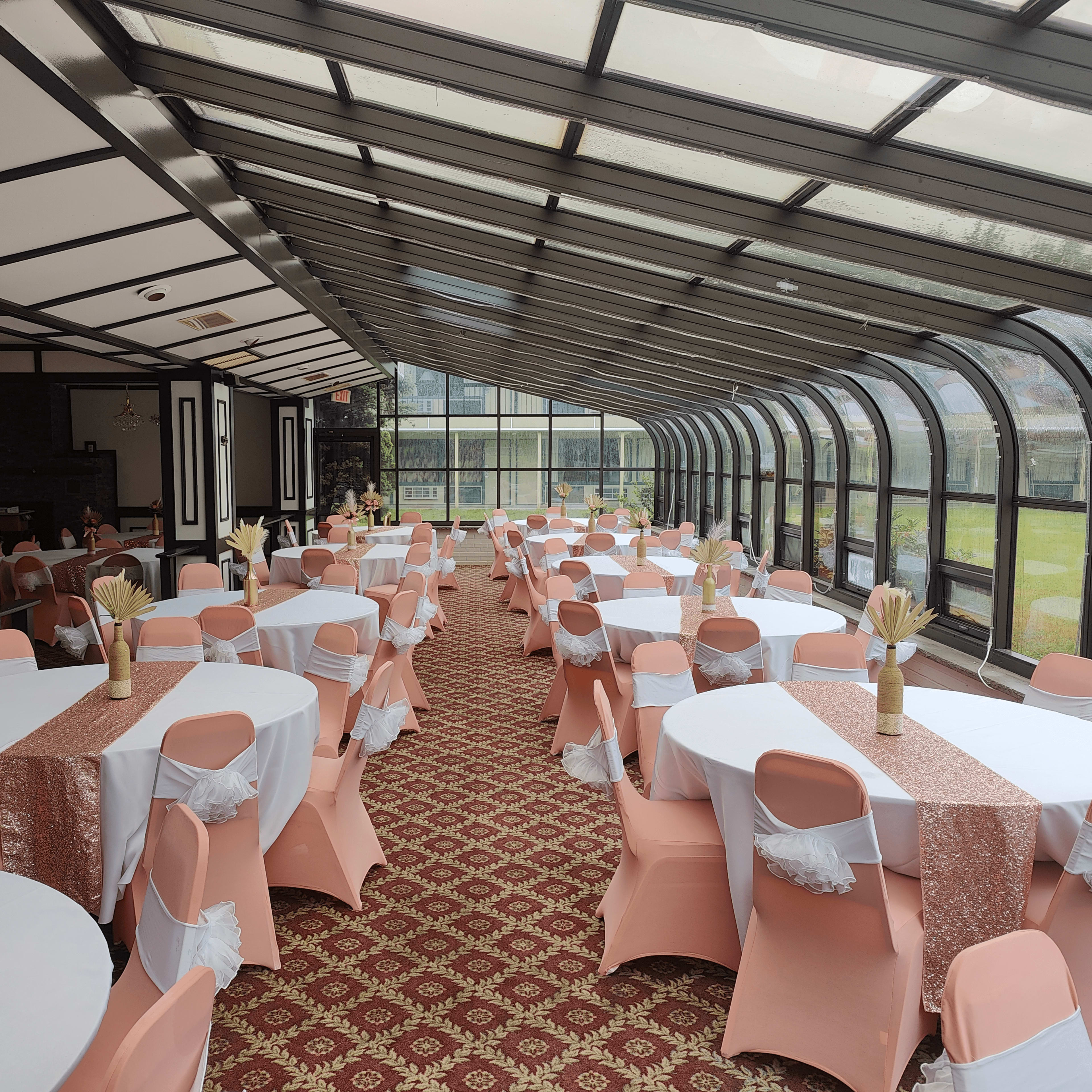 A banquet hall with round tables covered in white cloths and pink chair covers, arranged under a glass roof with large windows overlooking a green area.
