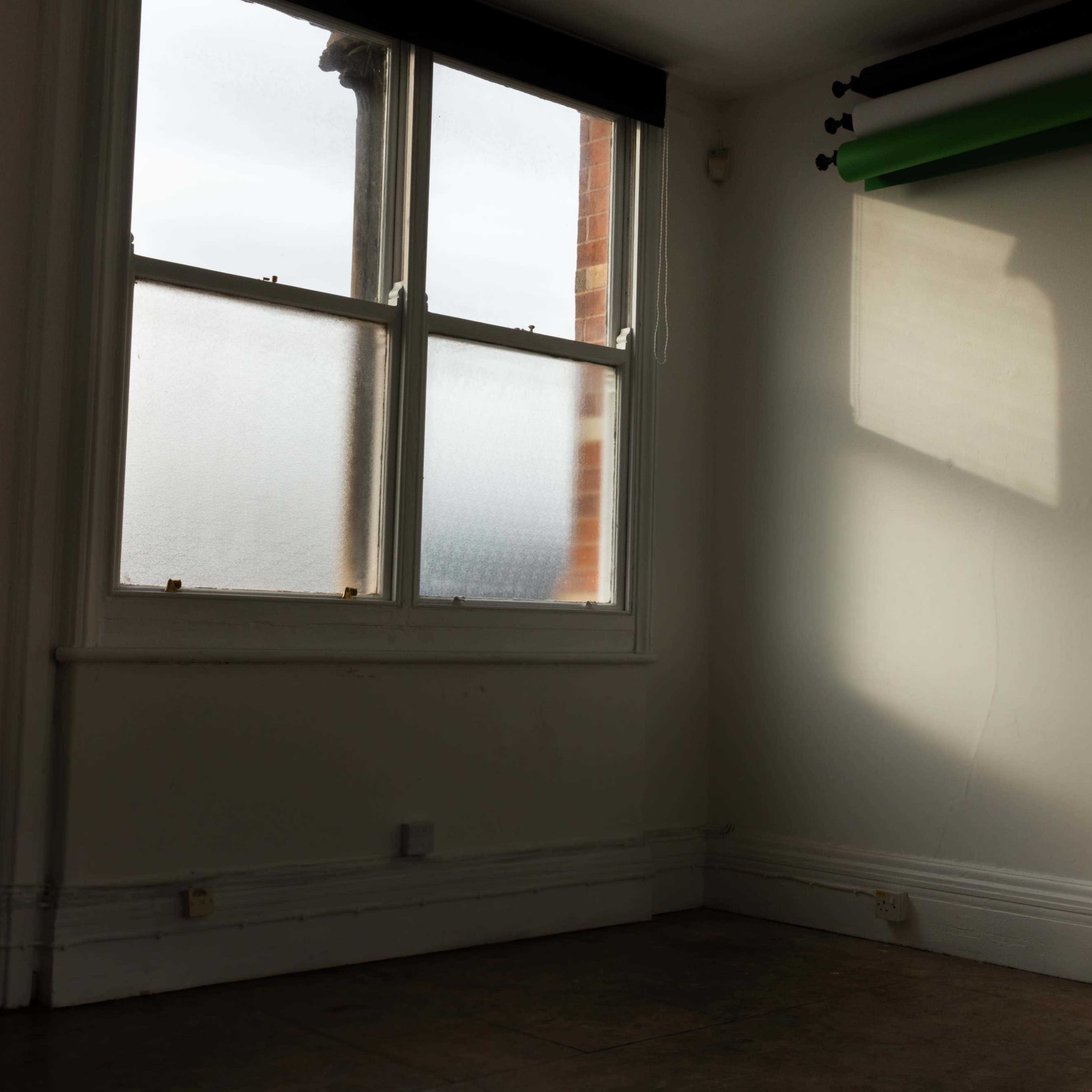 A sunlit room with a large window and a green backdrop roll hanging on the wall.
