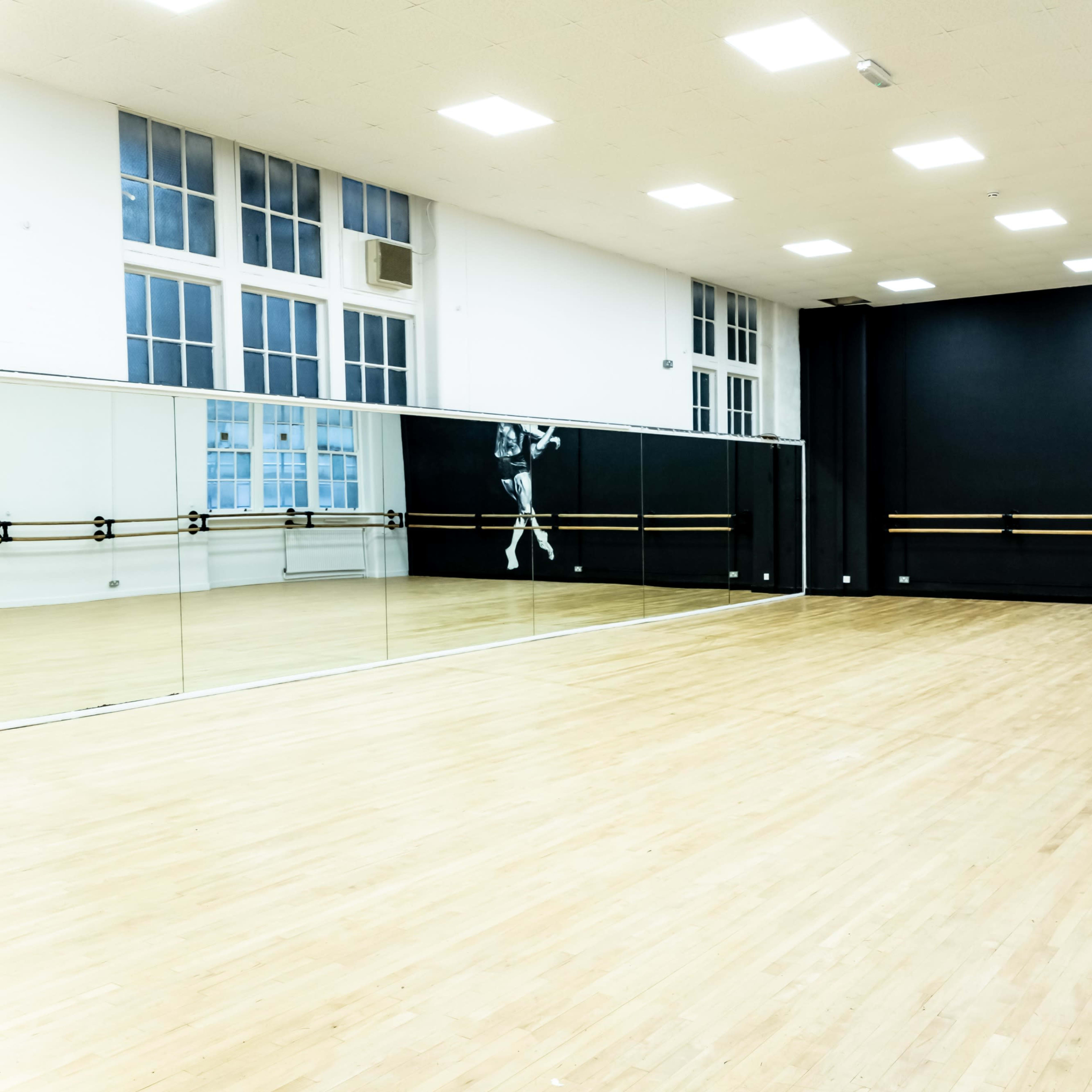A spacious, brightly lit dance studio with wooden flooring, large mirrors, and a black wall featuring a mural of a dancer.