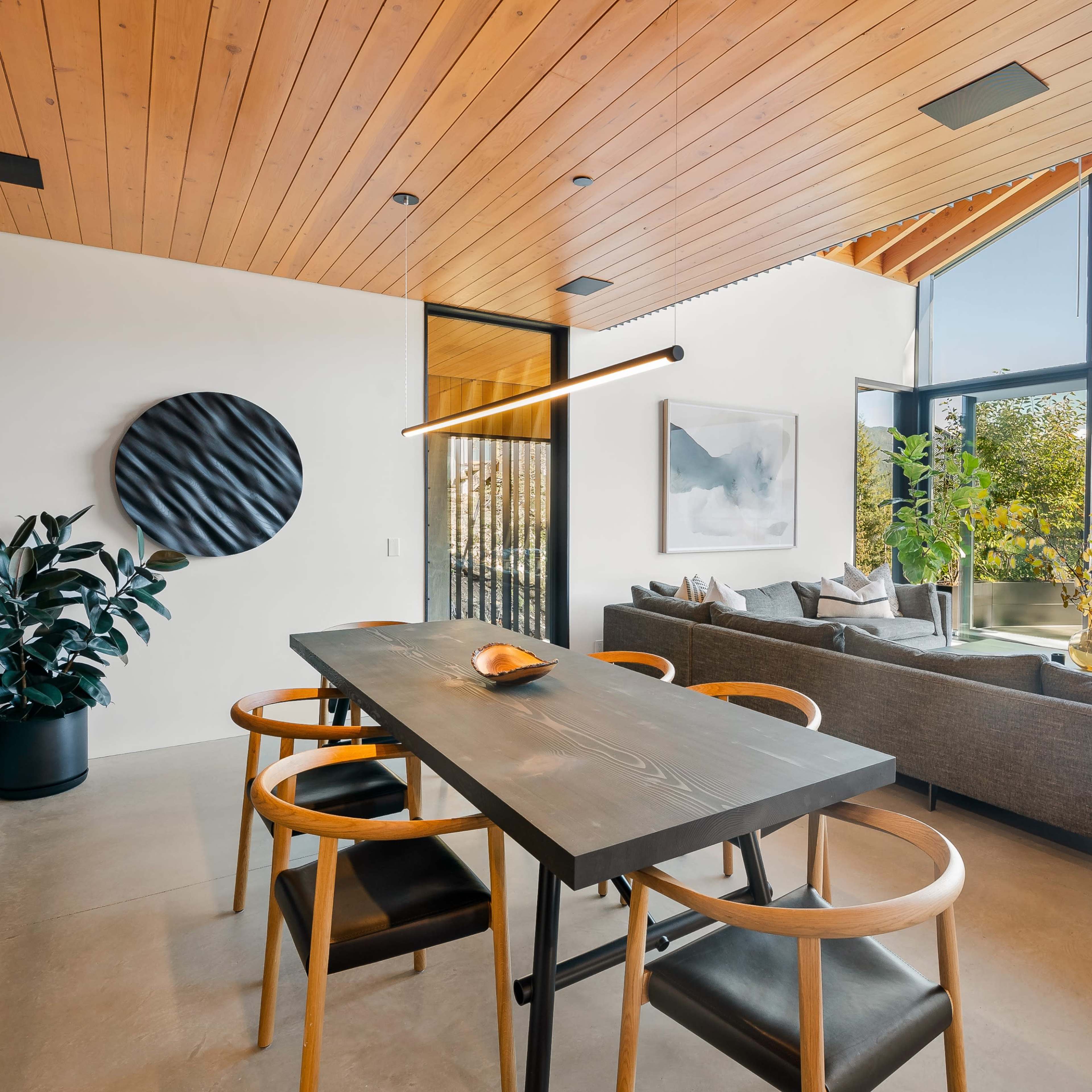 A modern dining area features a black table with wooden chairs, surrounded by large windows that overlook a scenic outdoor space.