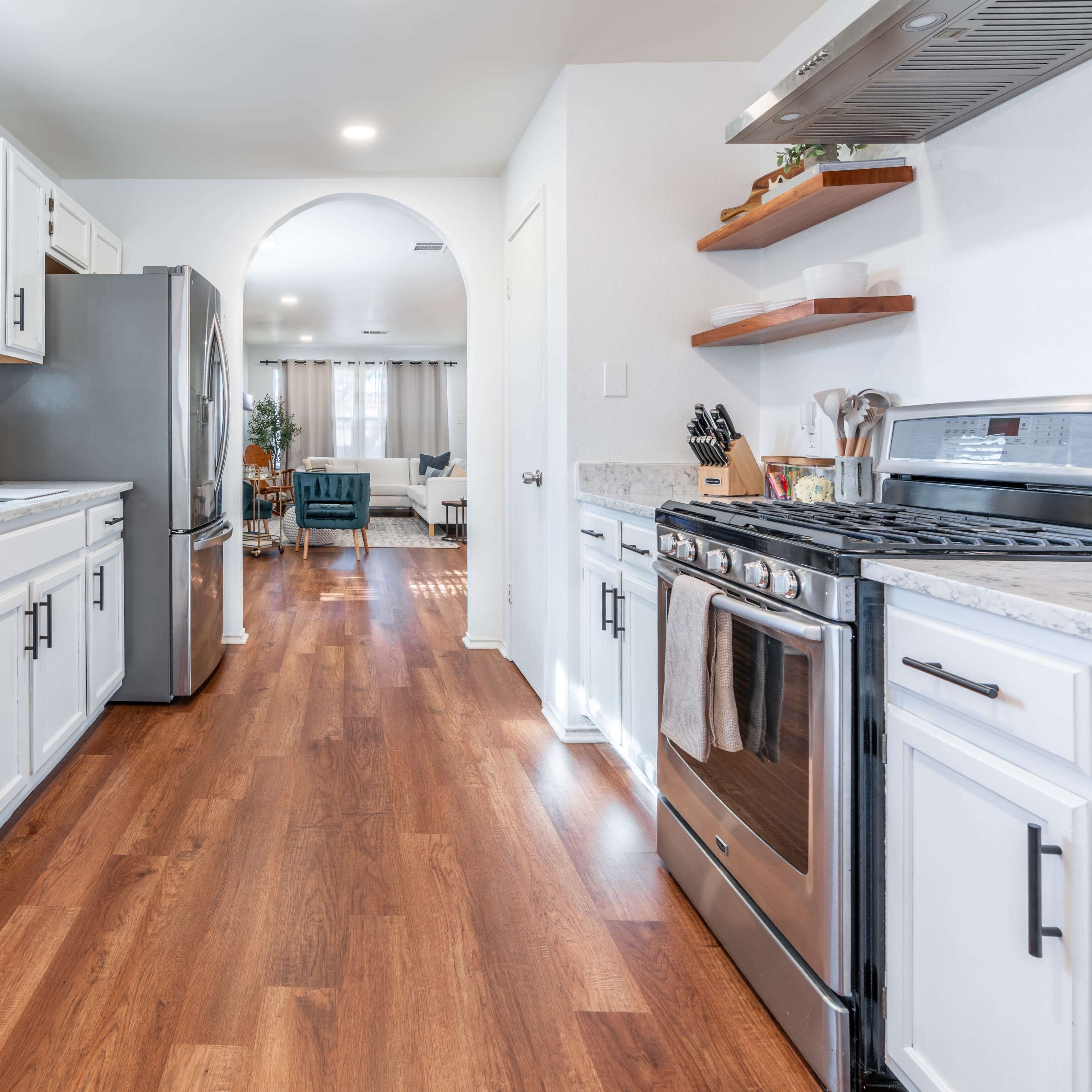 A modern kitchen features stainless steel appliances, white cabinetry, and hardwood flooring, with an open archway leading to a living area.