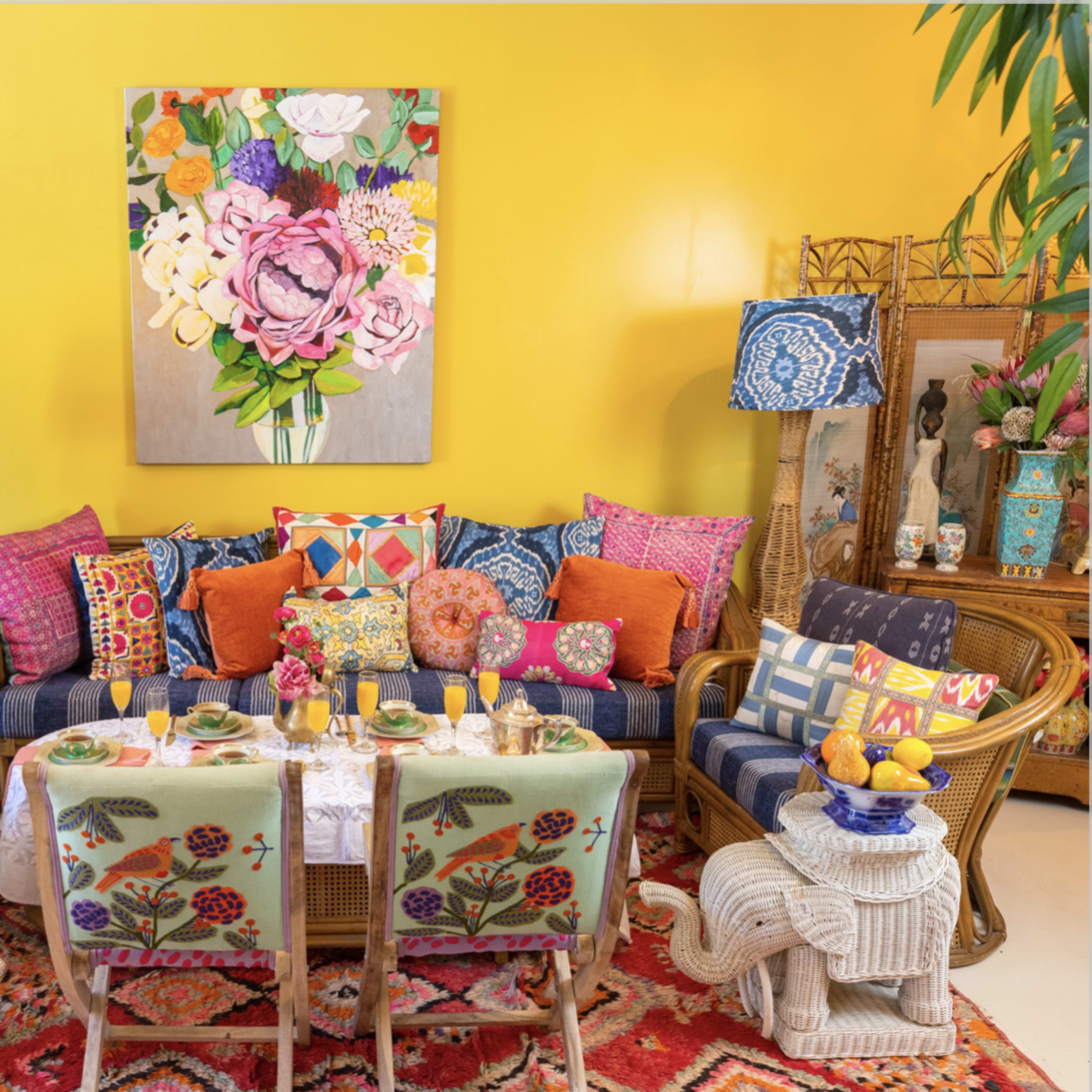 A brightly decorated living room features a patterned sofa surrounded by vibrant cushions and a dining table set with glasses and a floral centerpiece against a yellow wall.