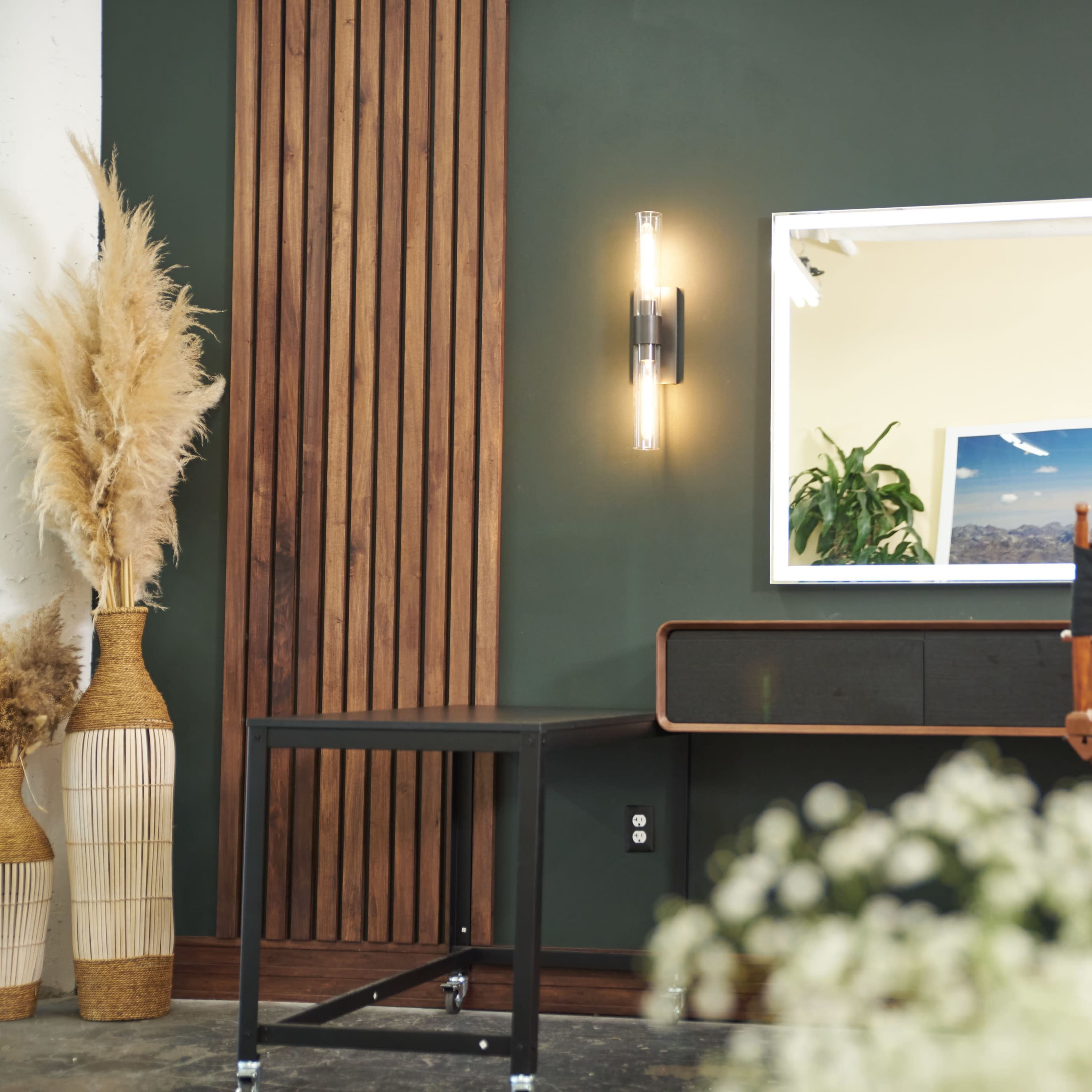 A minimalist interior design with a dark green wall, wooden accents, a lighted mirror, and a director's chair beside a table and decorative plants.