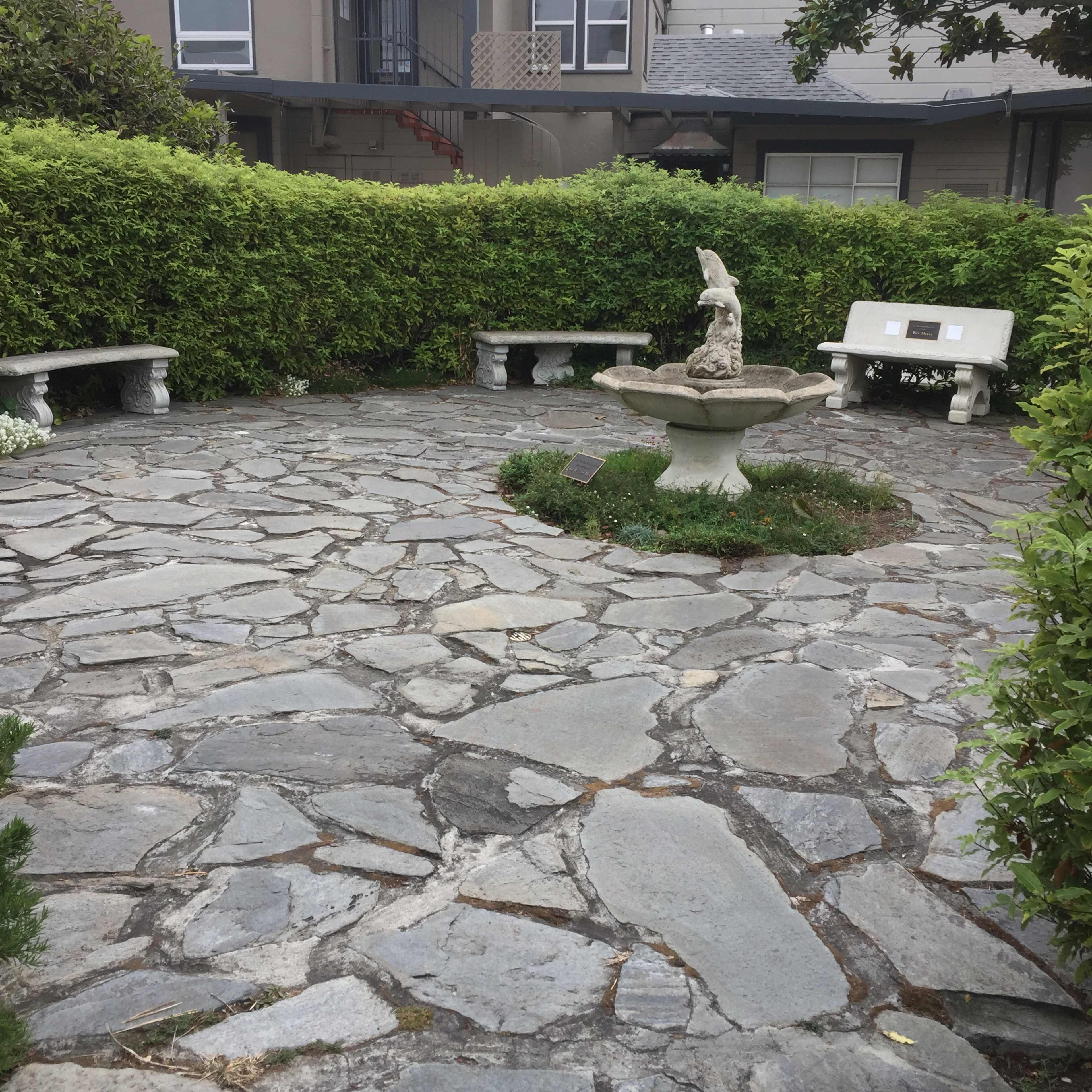 A stone-paved courtyard features a central fountain and stone benches surrounded by lush greenery.