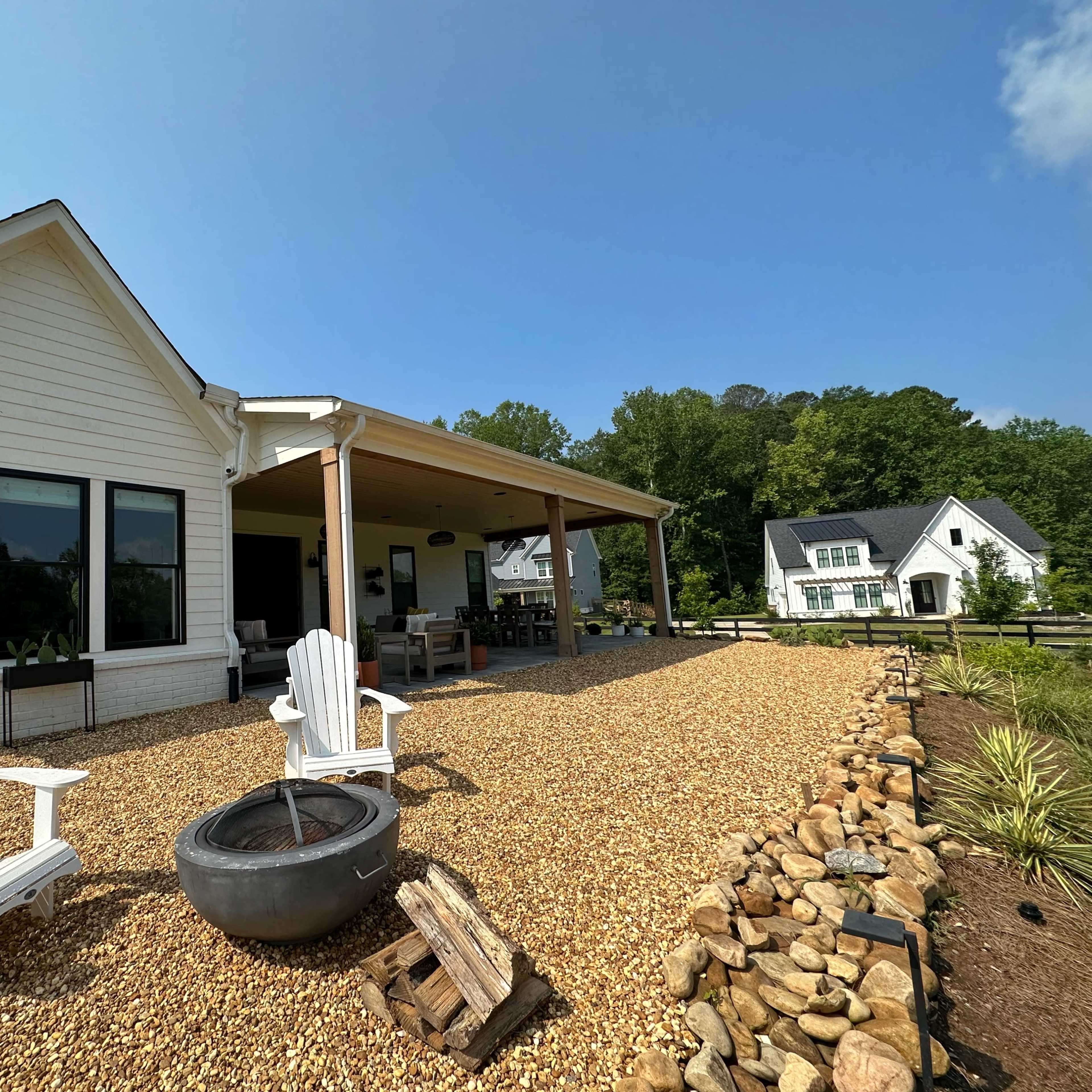 A white house with a covered patio and a landscaped outdoor area featuring gravel, chairs, and a fire pit.