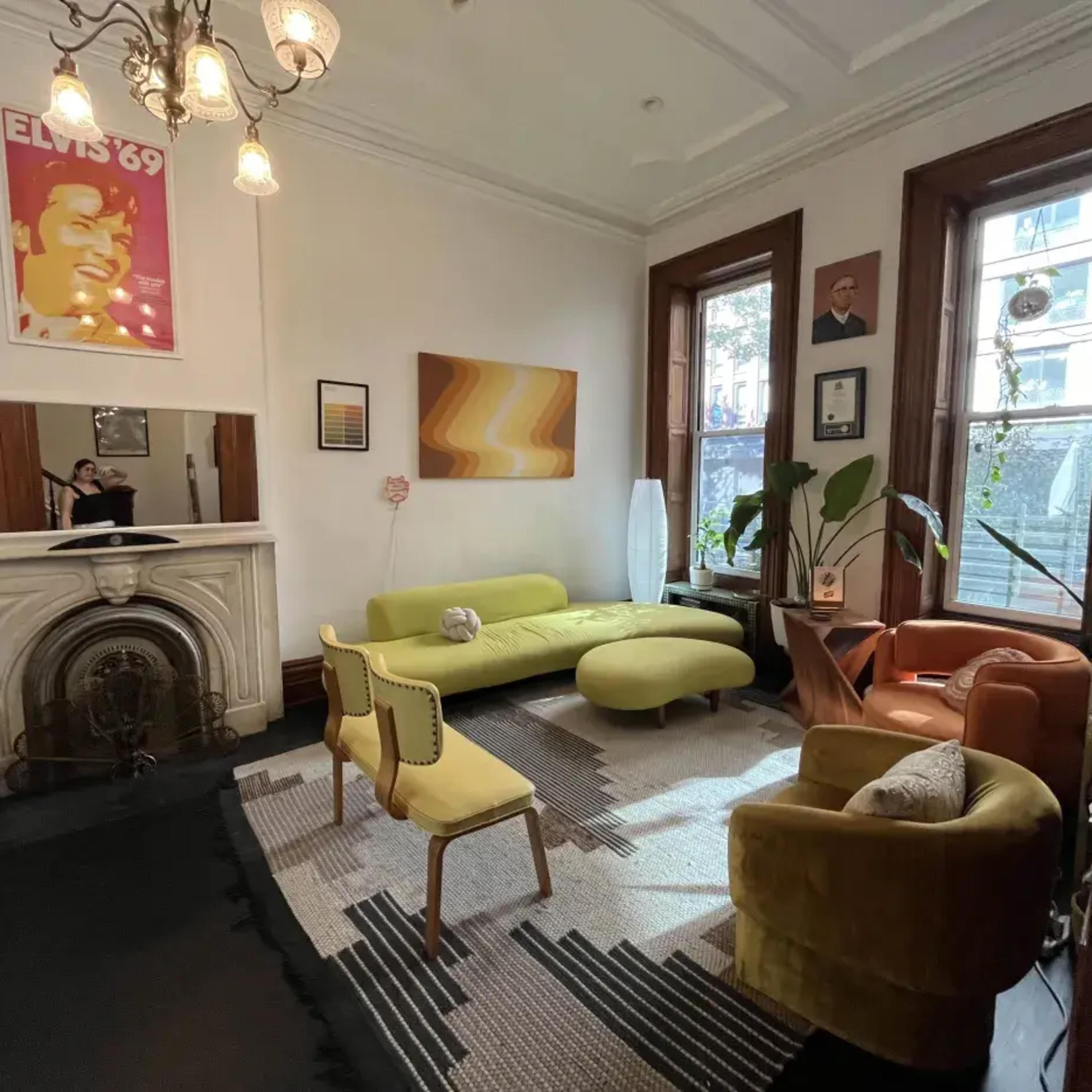 A stylish living room featuring a green sofa, a yellow chair, and a decorative fireplace, with large windows letting in natural light.