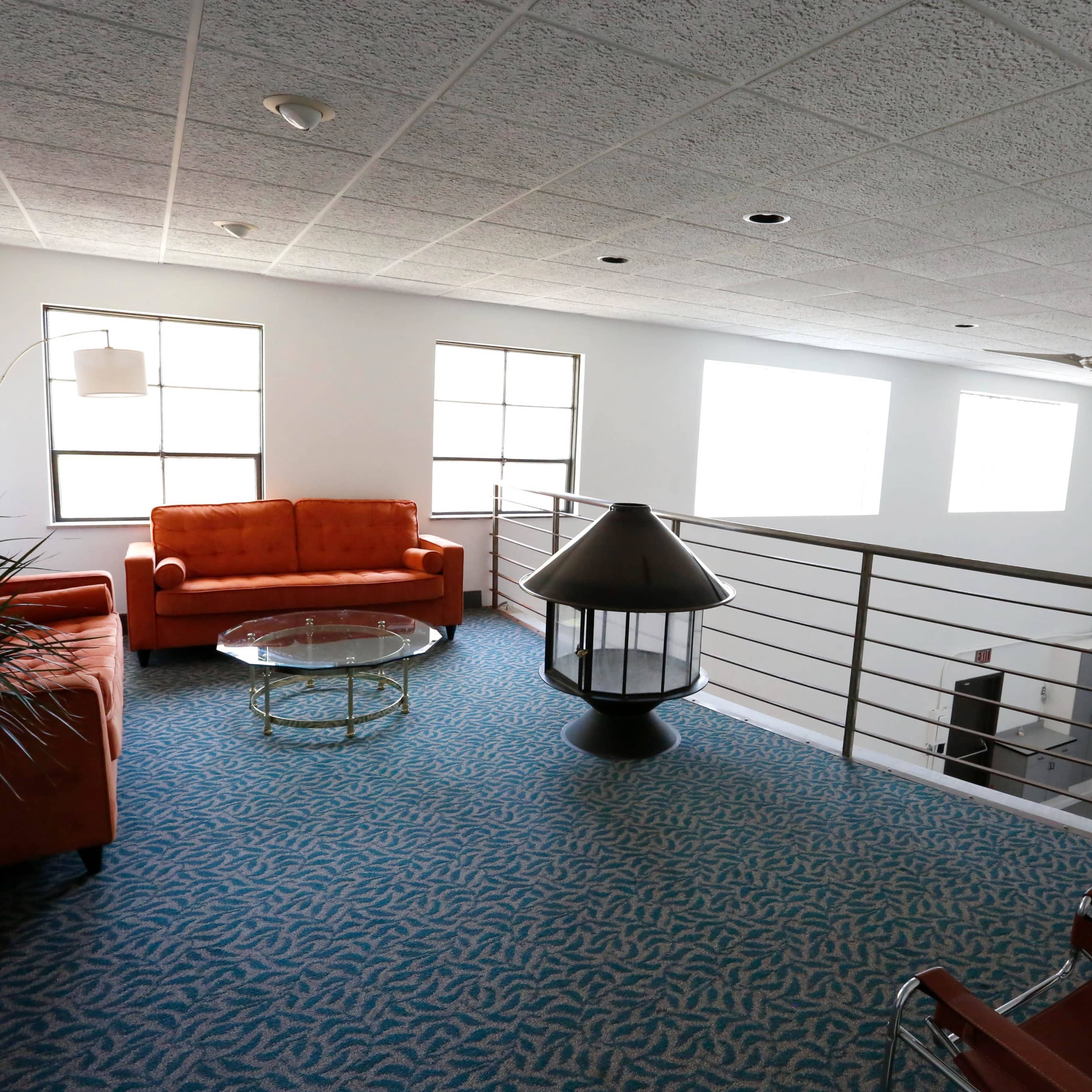 A loft-style living area with an orange couch, a glass coffee table, and a decorative lamp, surrounded by large windows and a railing.