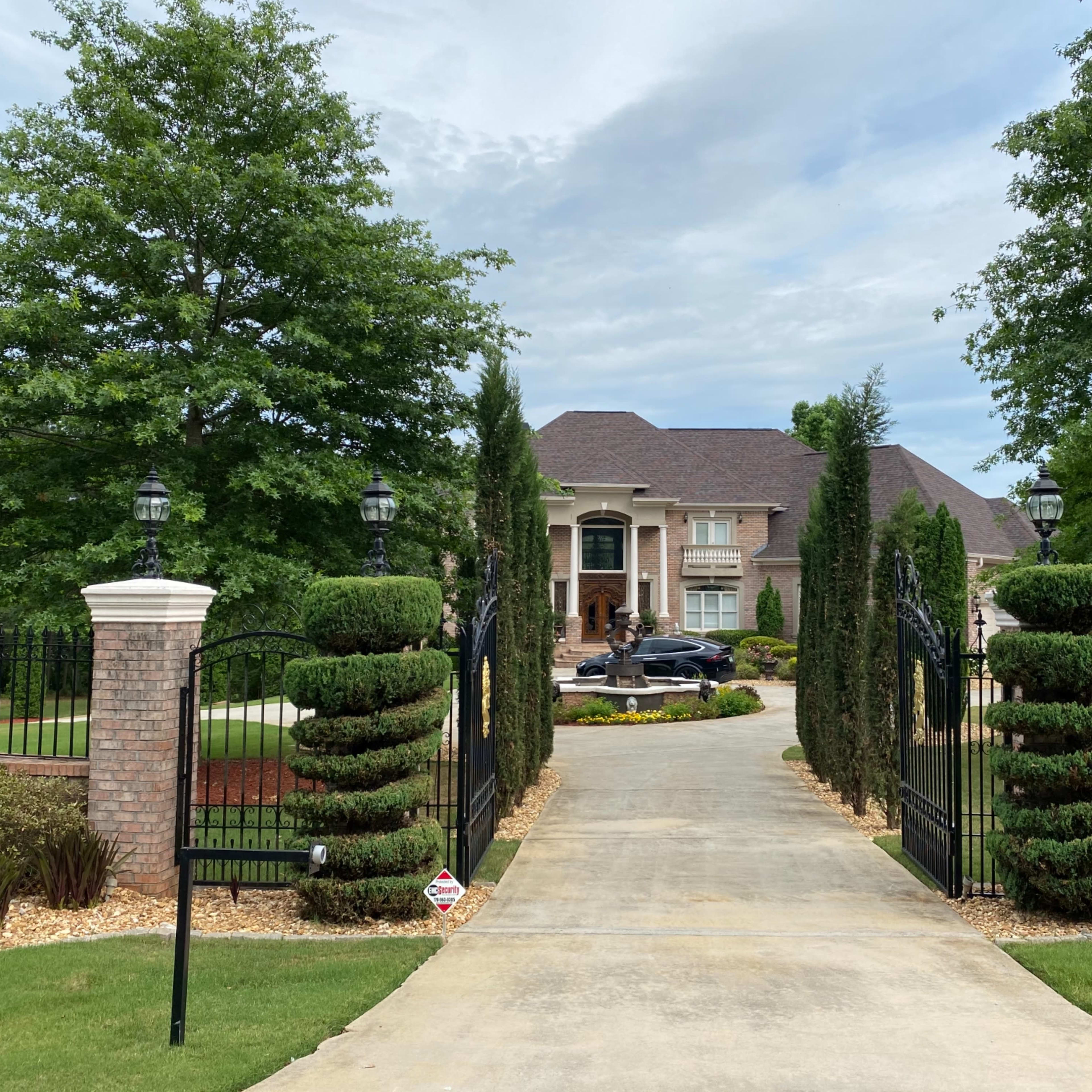 A long driveway leads to a large brick house surrounded by neatly trimmed trees and manicured landscaping.