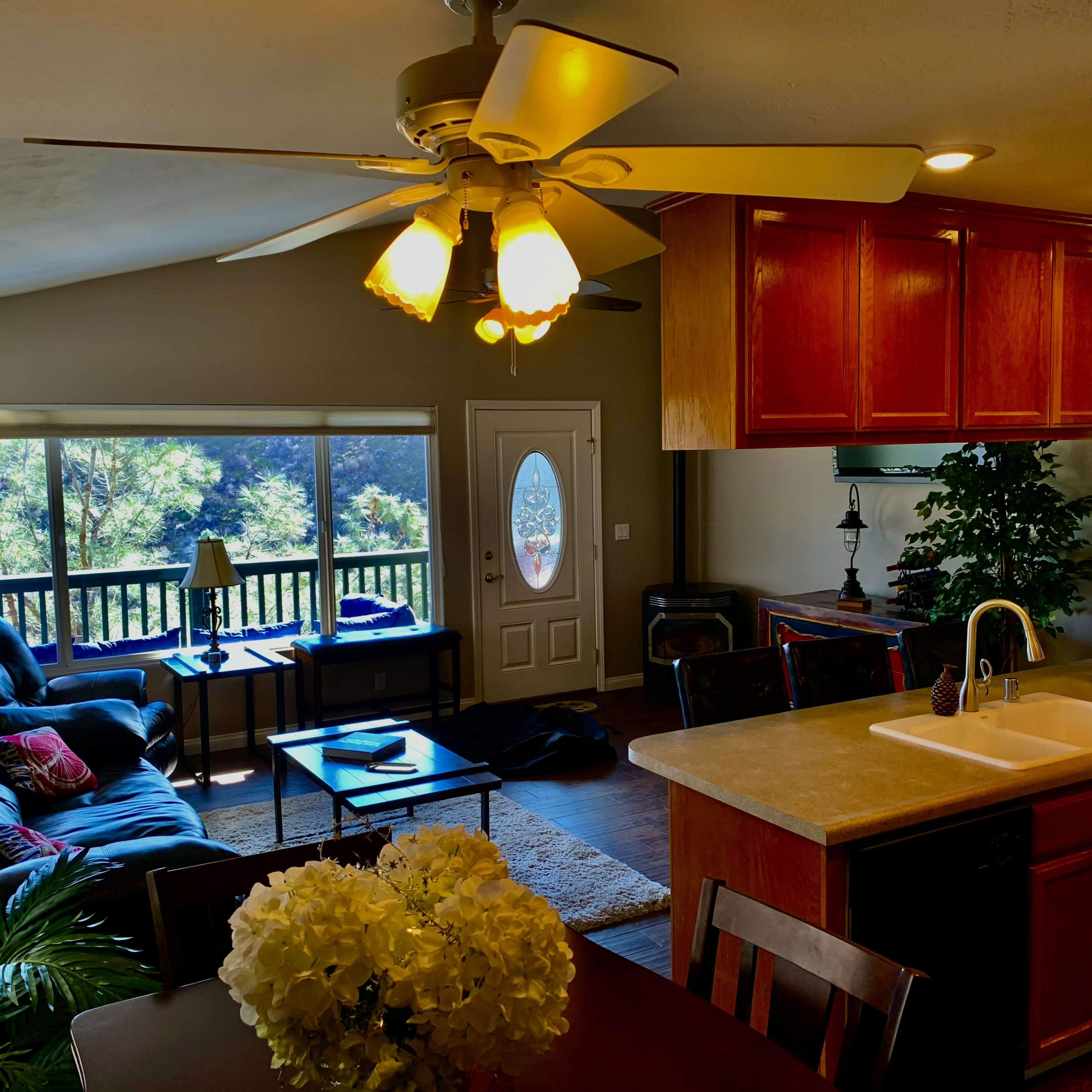 A cozy living room with a large window, a dark leather sofa, a wooden dining table, and a ceiling fan overhead.
