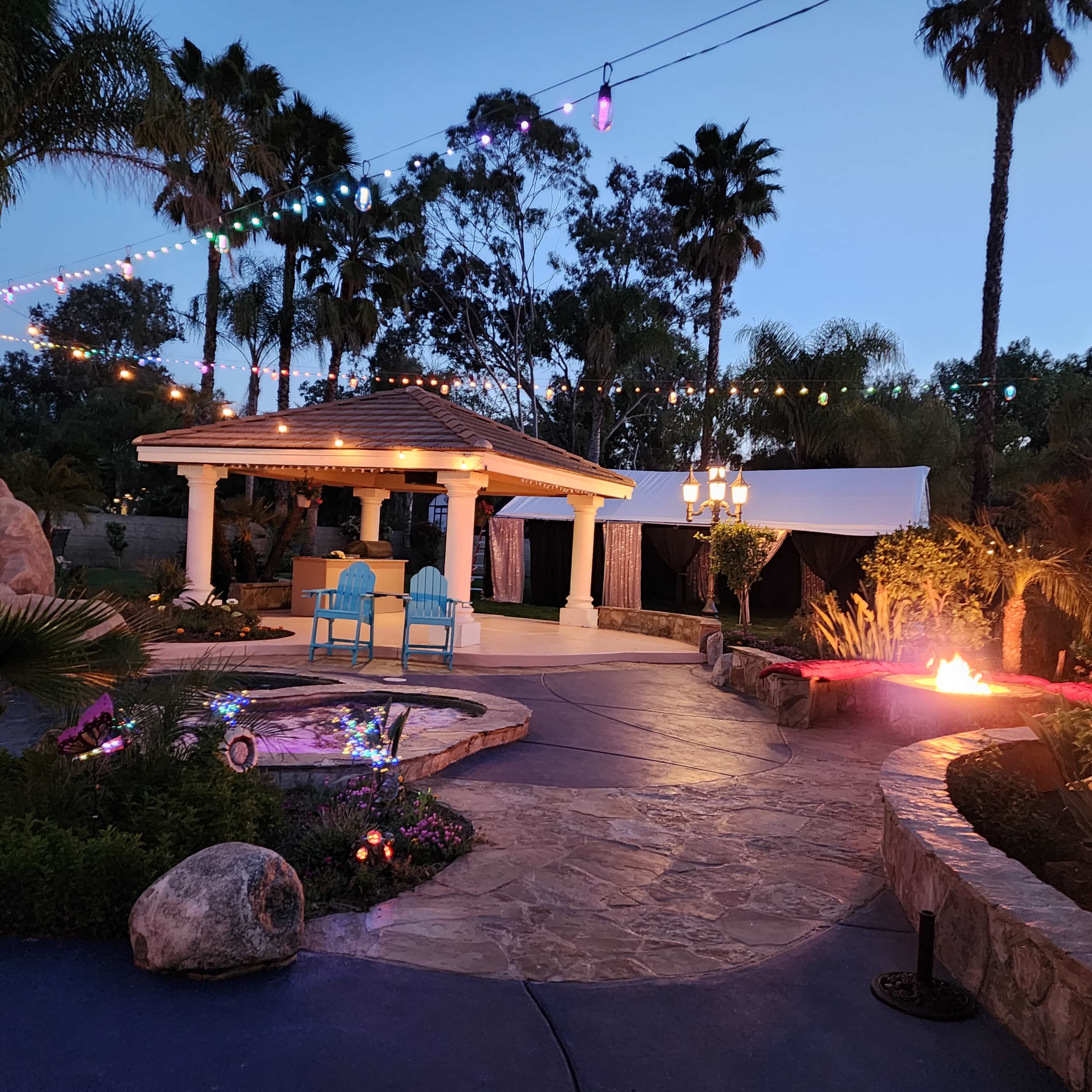 A landscaped outdoor area featuring a gazebo, two blue chairs, and decorative lighting amidst palm trees and a fire pit.