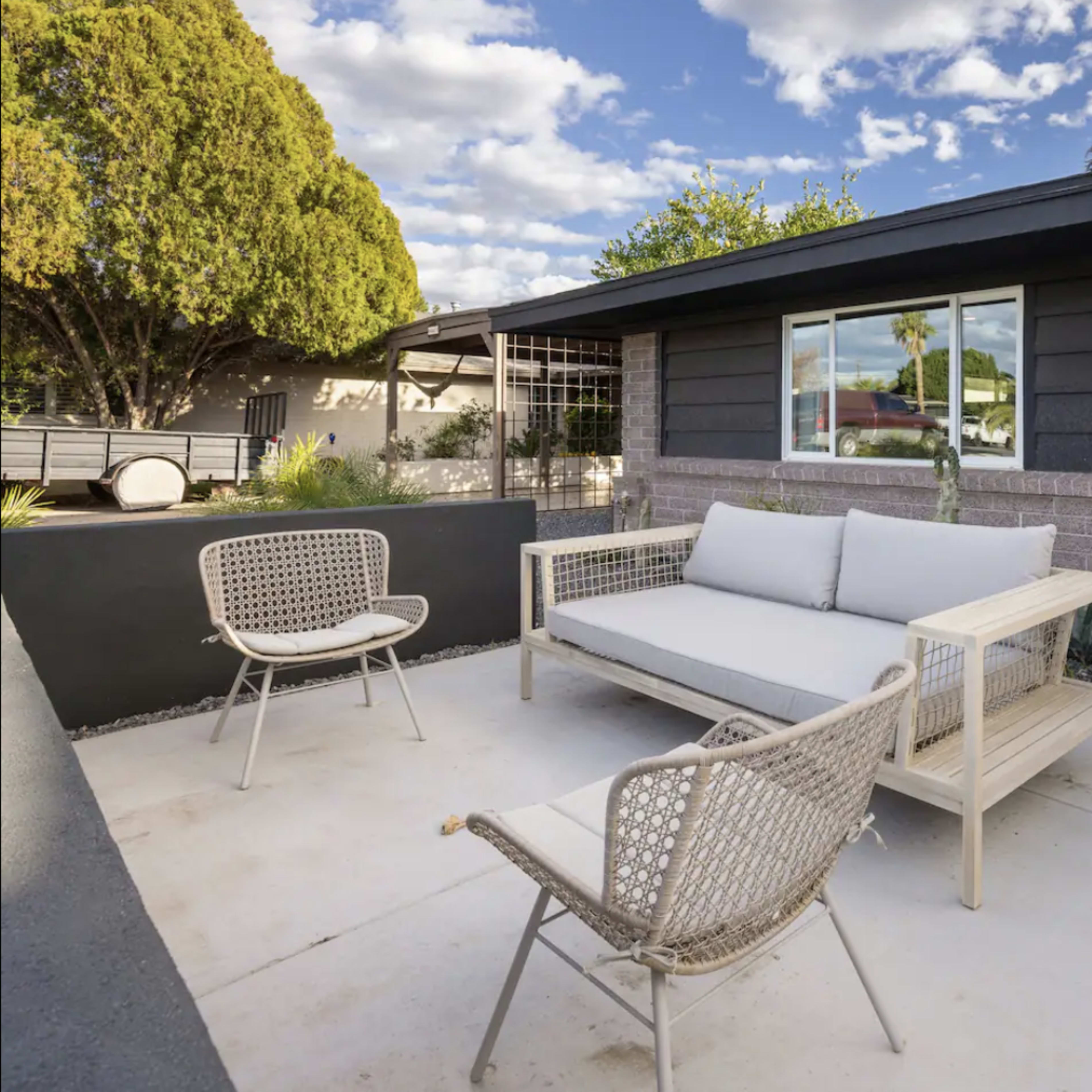 A modern outdoor seating area features a light-colored sofa and two woven chairs on a concrete patio, surrounded by low landscaping and a house with dark siding.