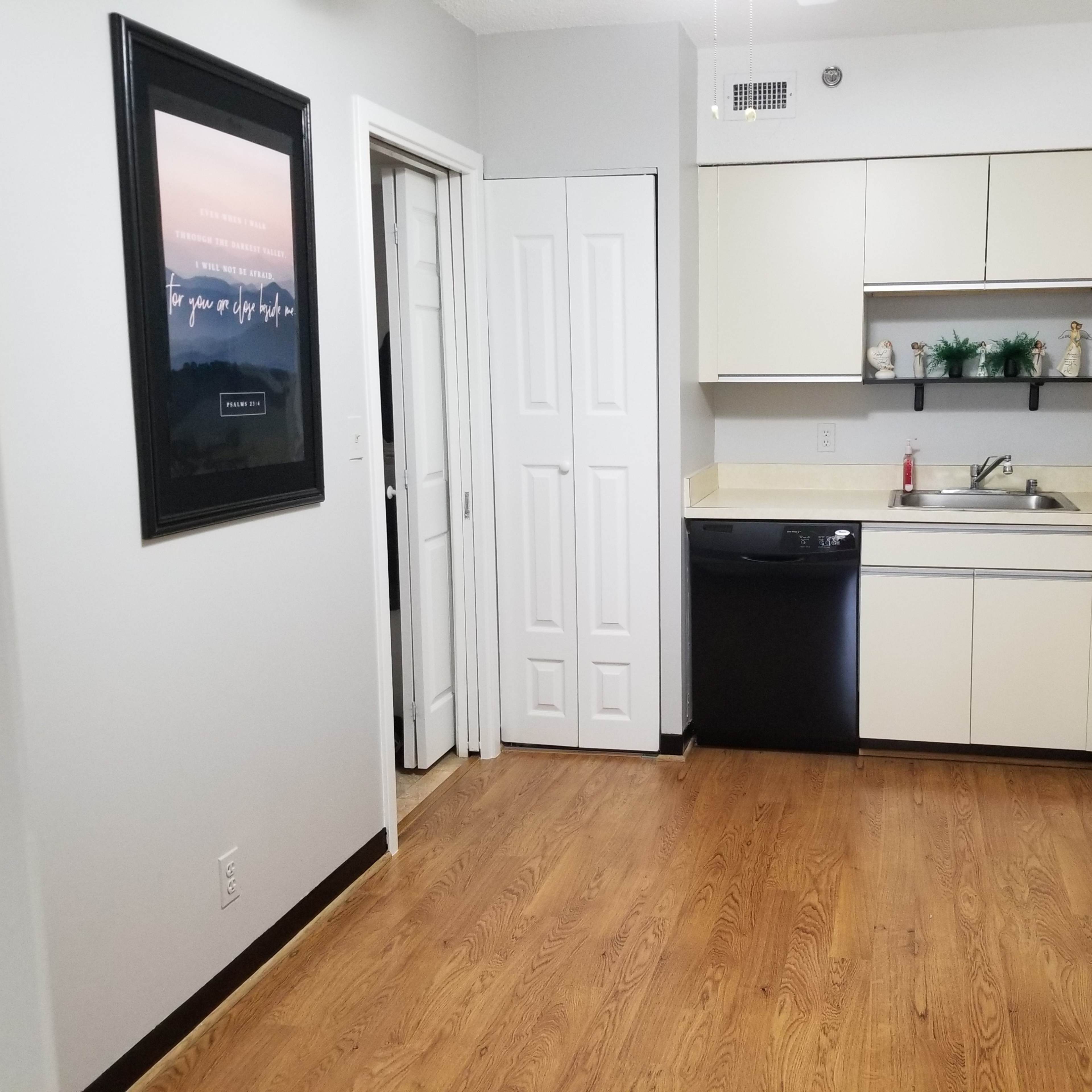 A clean kitchen with light-colored cabinets, a black dishwasher, an oven, and a small dining area against a wall with a framed picture.