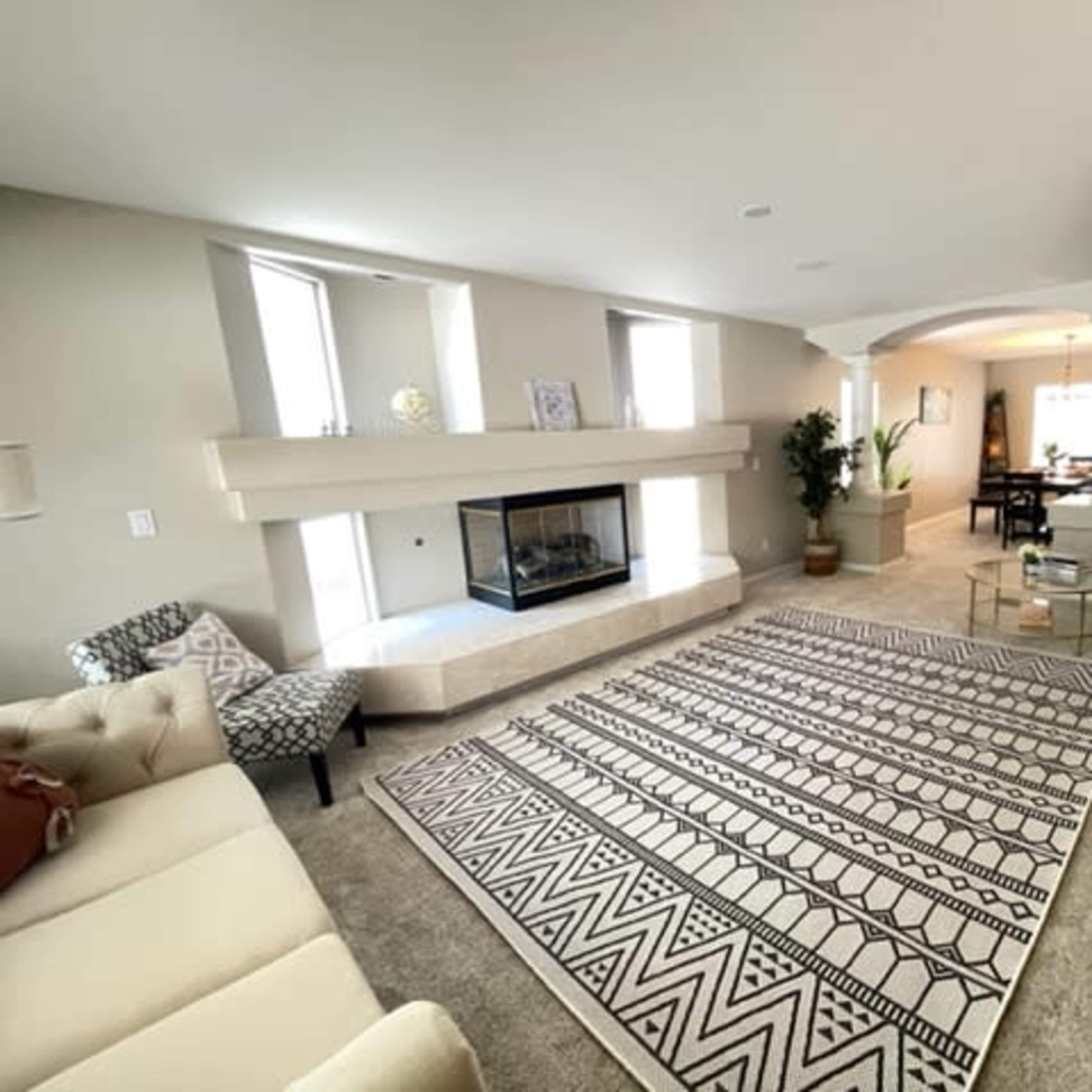 A spacious living room featuring a beige sectional sofa, a modern fireplace, a patterned area rug, and a small glass coffee table.