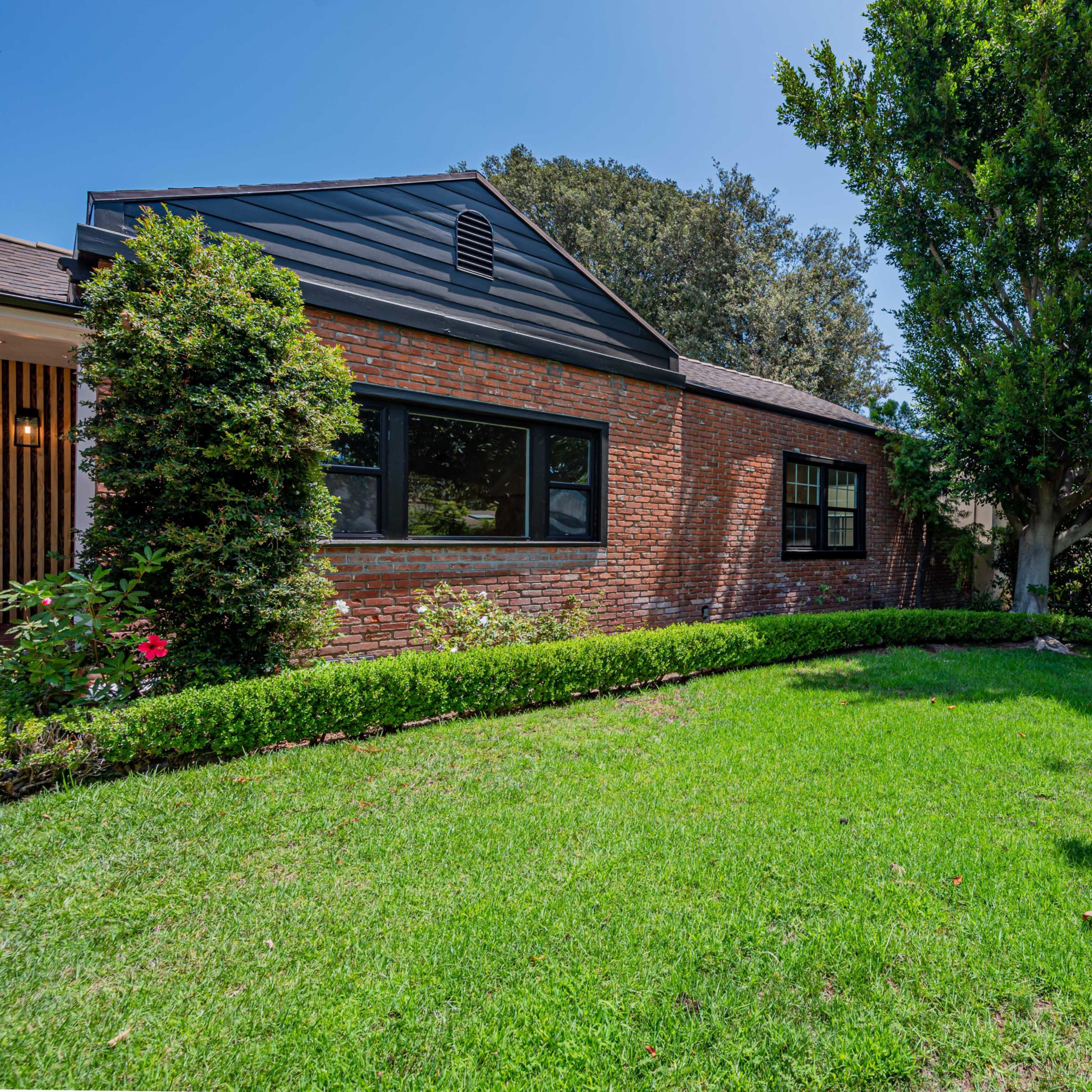 A brick house with black accents sits beside a manicured lawn surrounded by shrubs and trees.