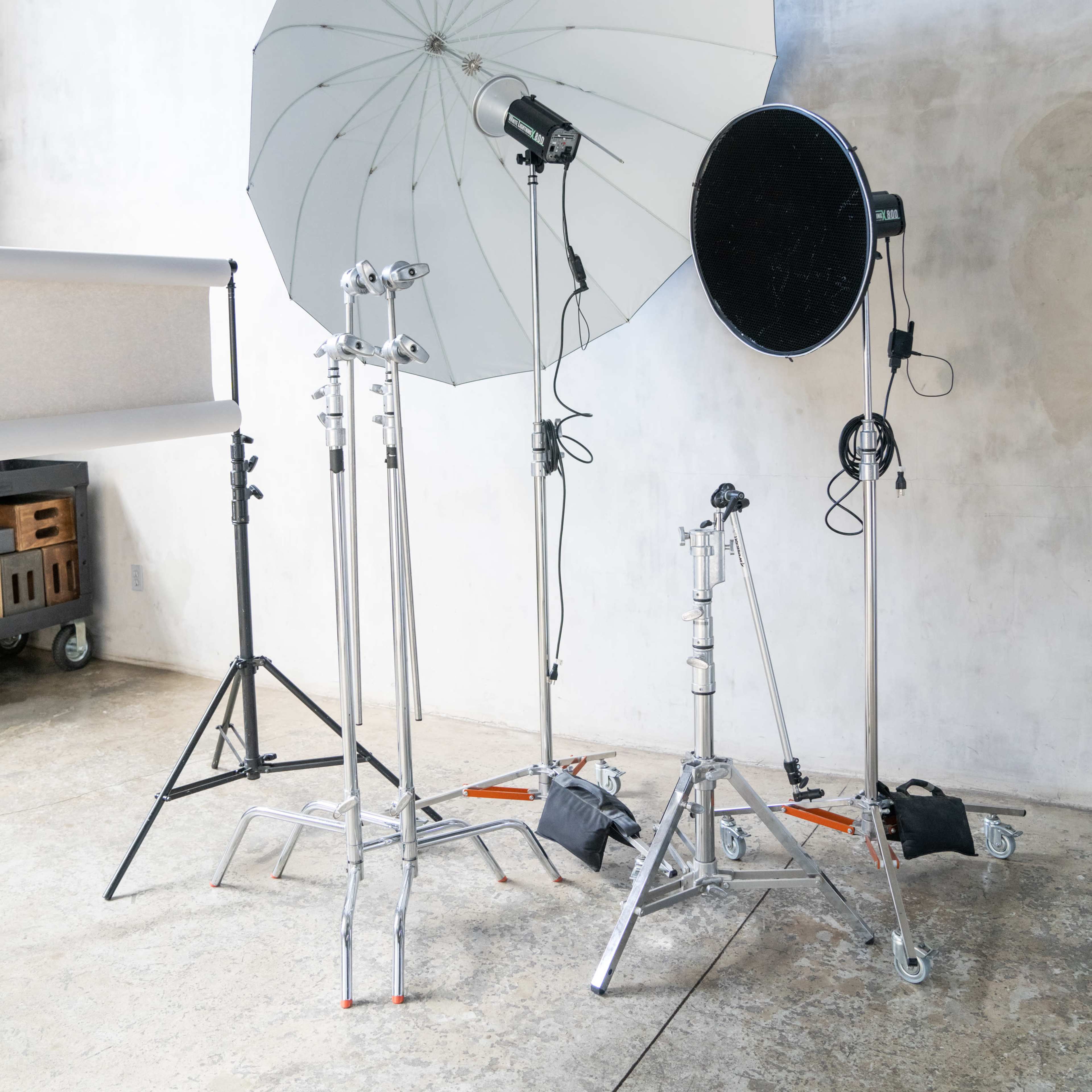 Various photography equipment, including light stands, a large umbrella reflector, and a circular reflector against a concrete wall.