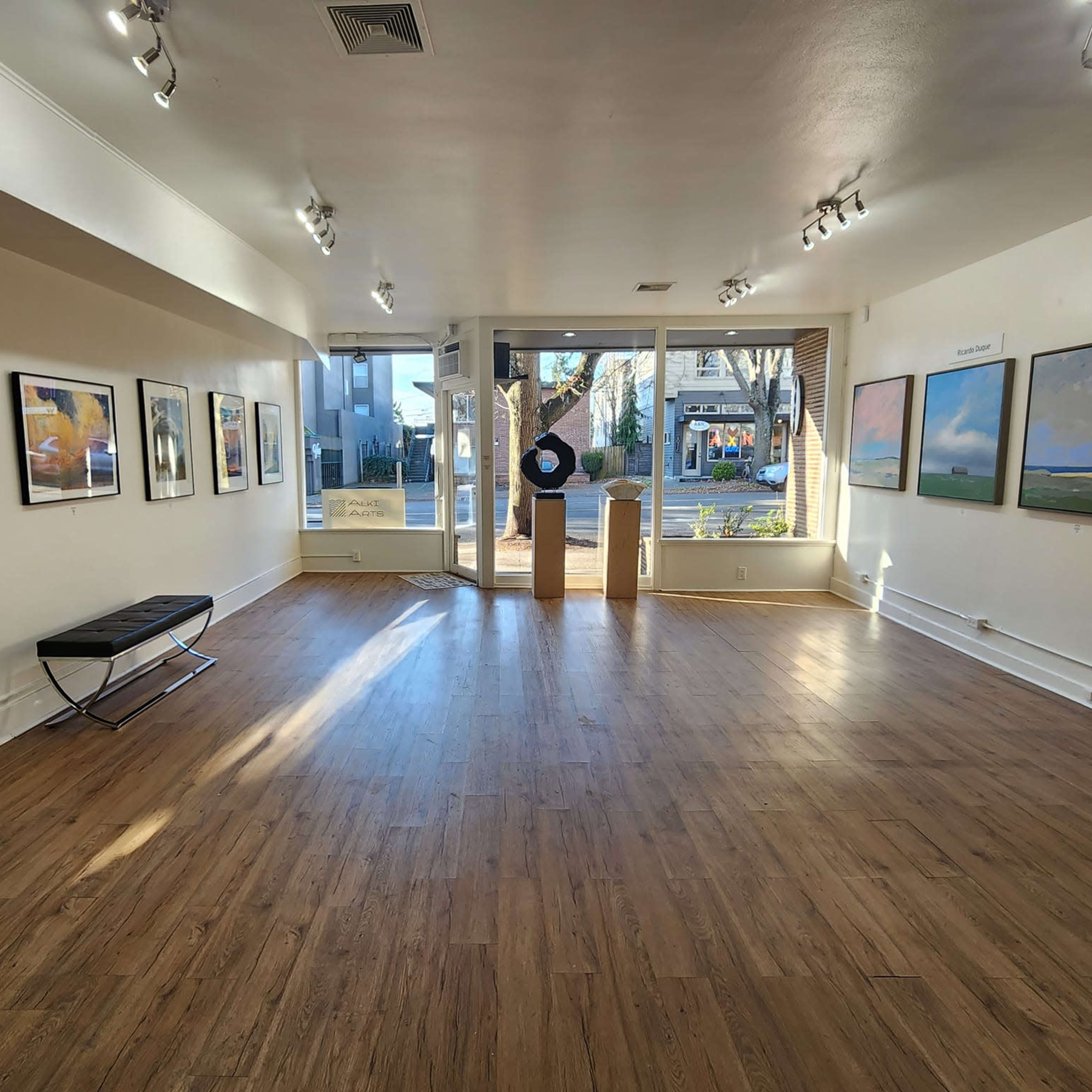 An art gallery with wooden flooring and several framed paintings displayed on the walls, leading to a large window at the entrance.