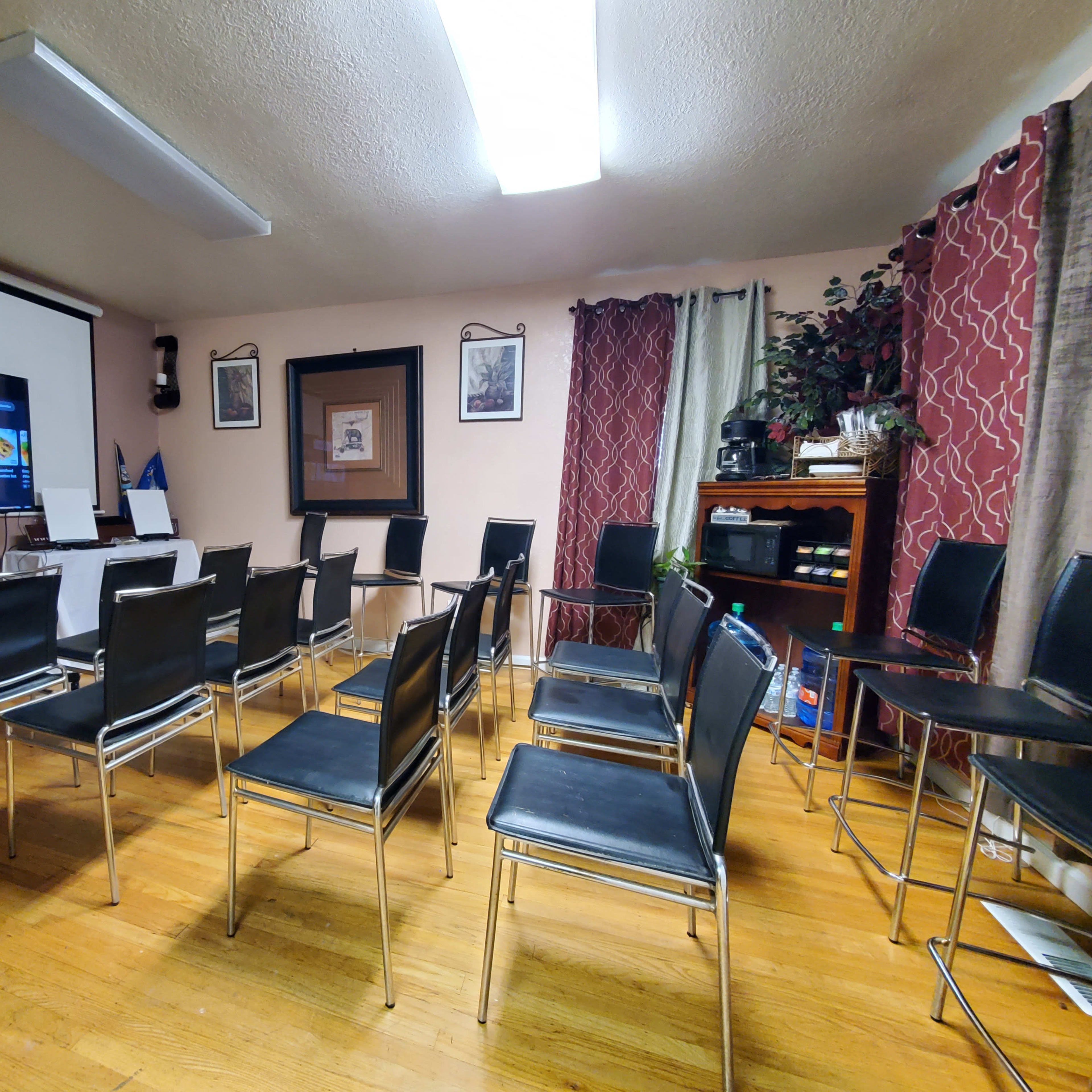 A room set up for a meeting or presentation features a screen displaying information, surrounded by empty black chairs arranged in a semi-circle.