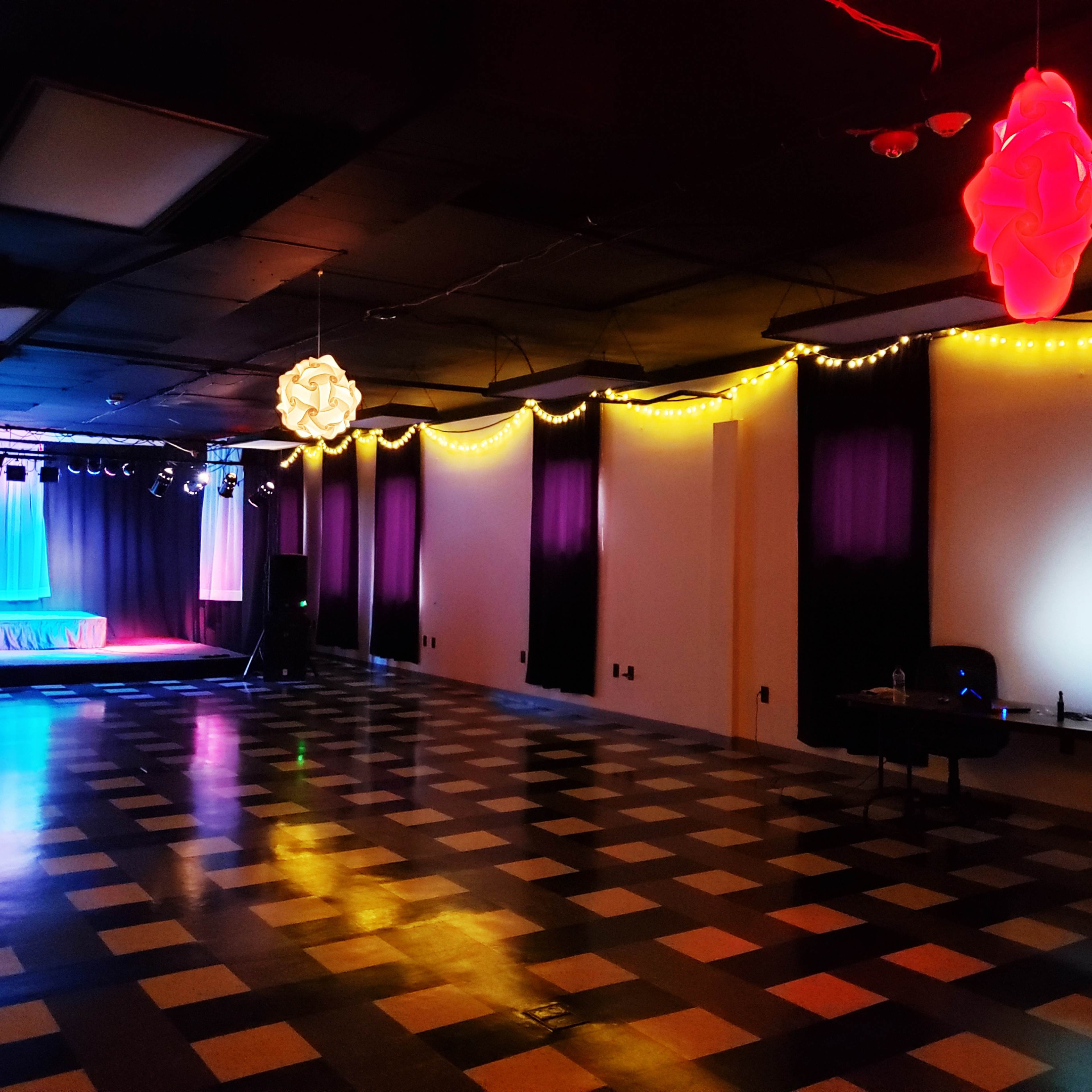 An empty event space with a stage in the background, decorated with colorful lighting and checkered flooring.