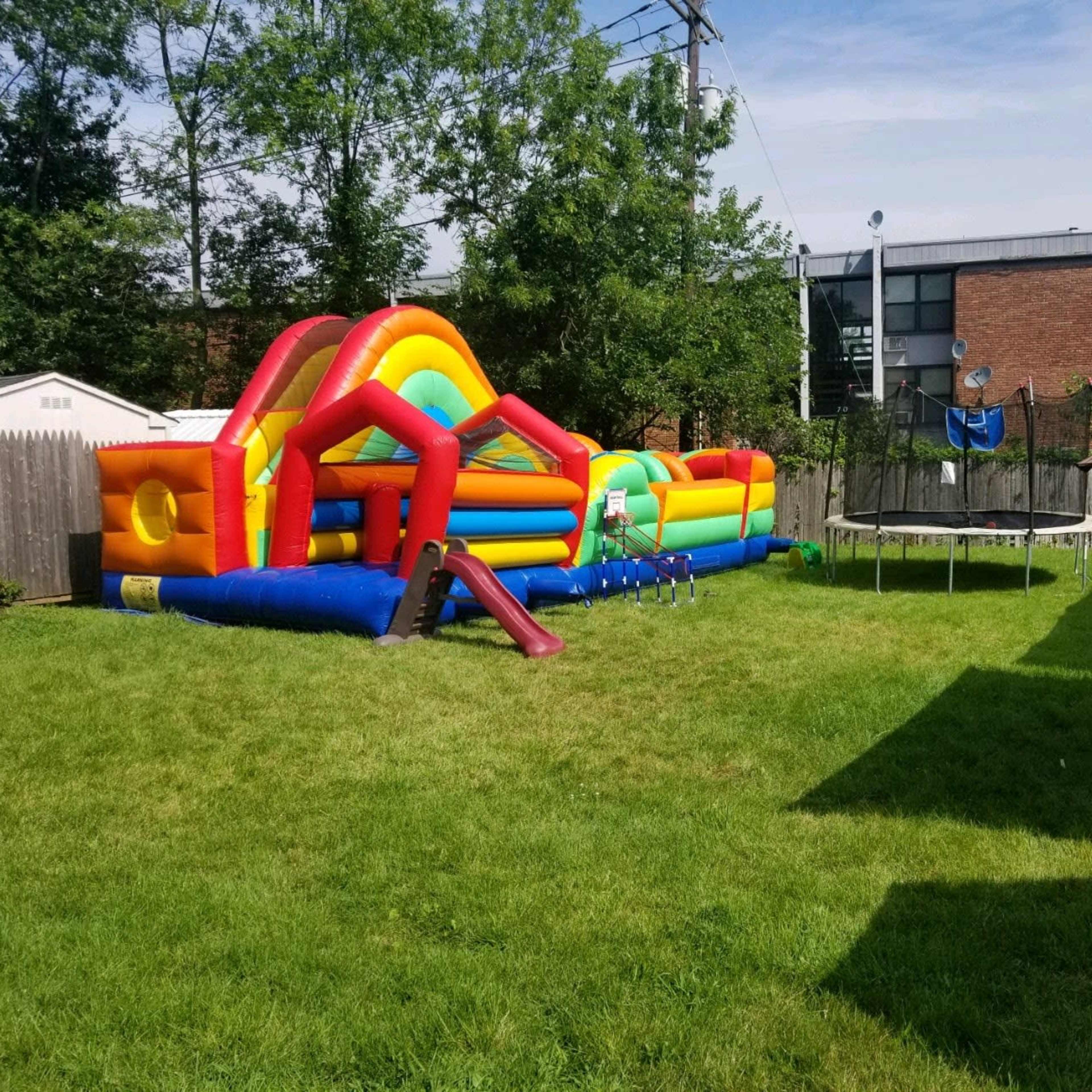 A backyard with a colorful inflatable bounce house and a trampoline near a grassy area with picnic tables set up under a canopy.