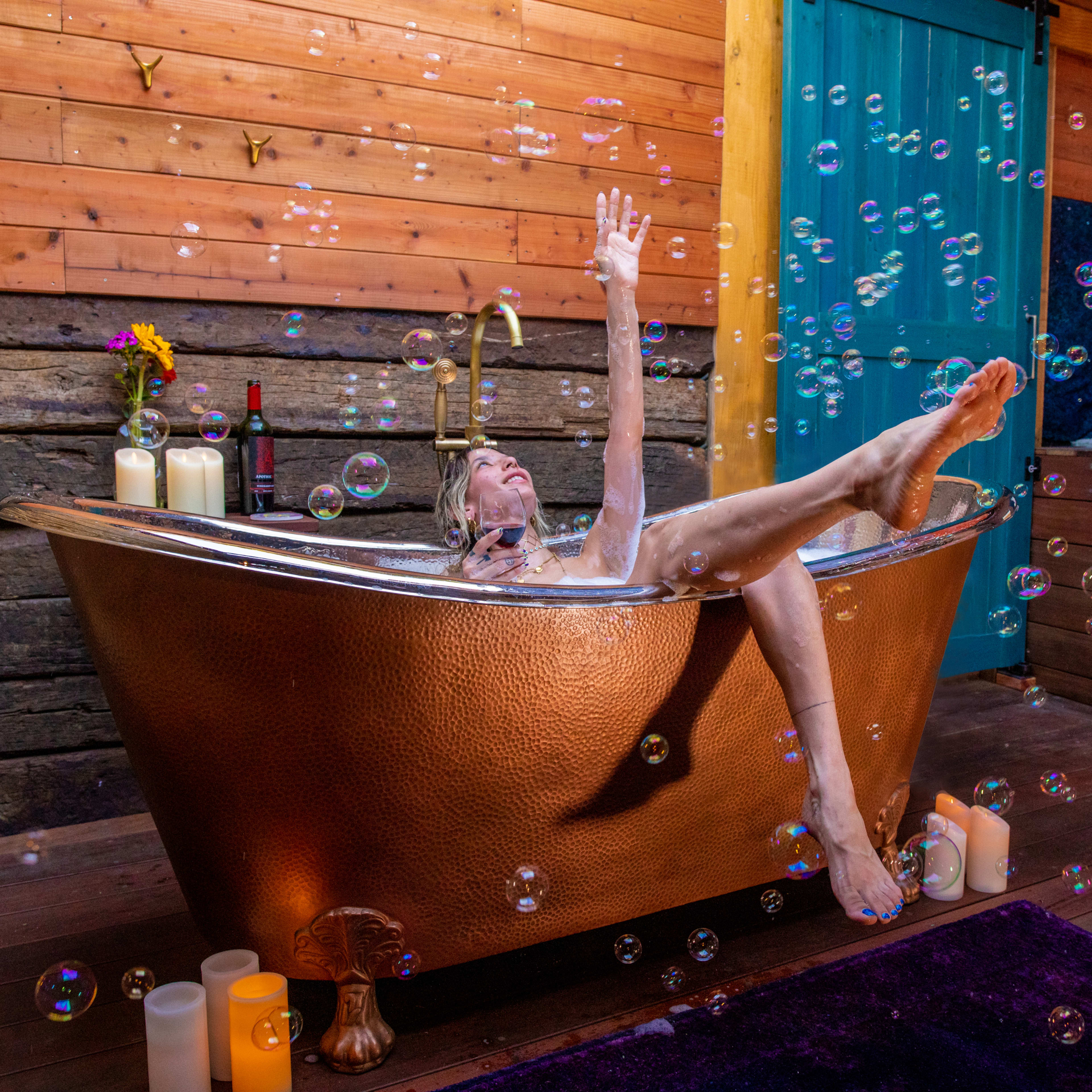 A person is reclining in a copper bathtub surrounded by bubbles and candles in a rustic wooden room.