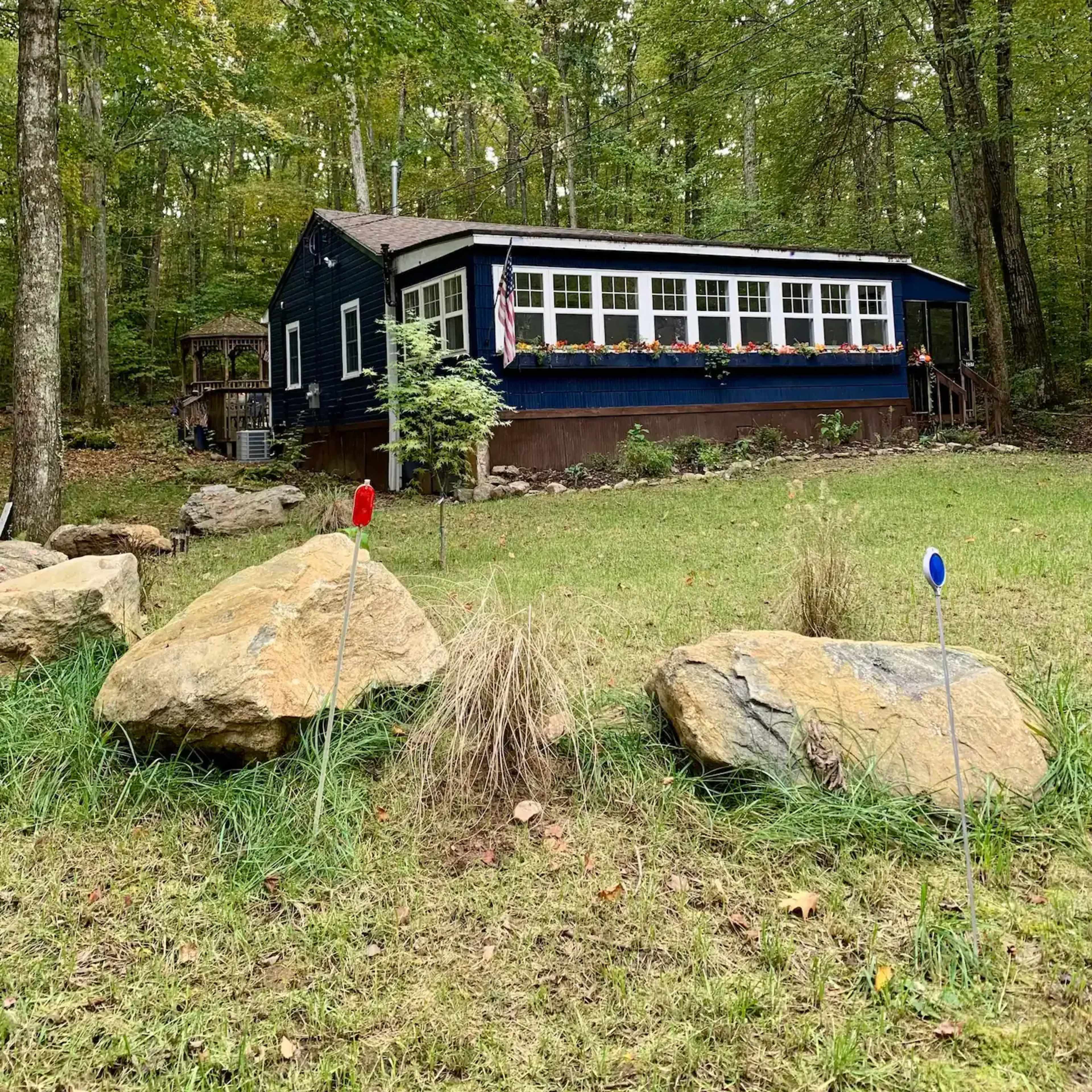 A blue house with a porch and large windows is nestled among trees in a grassy area, surrounded by boulders and decorative markers.