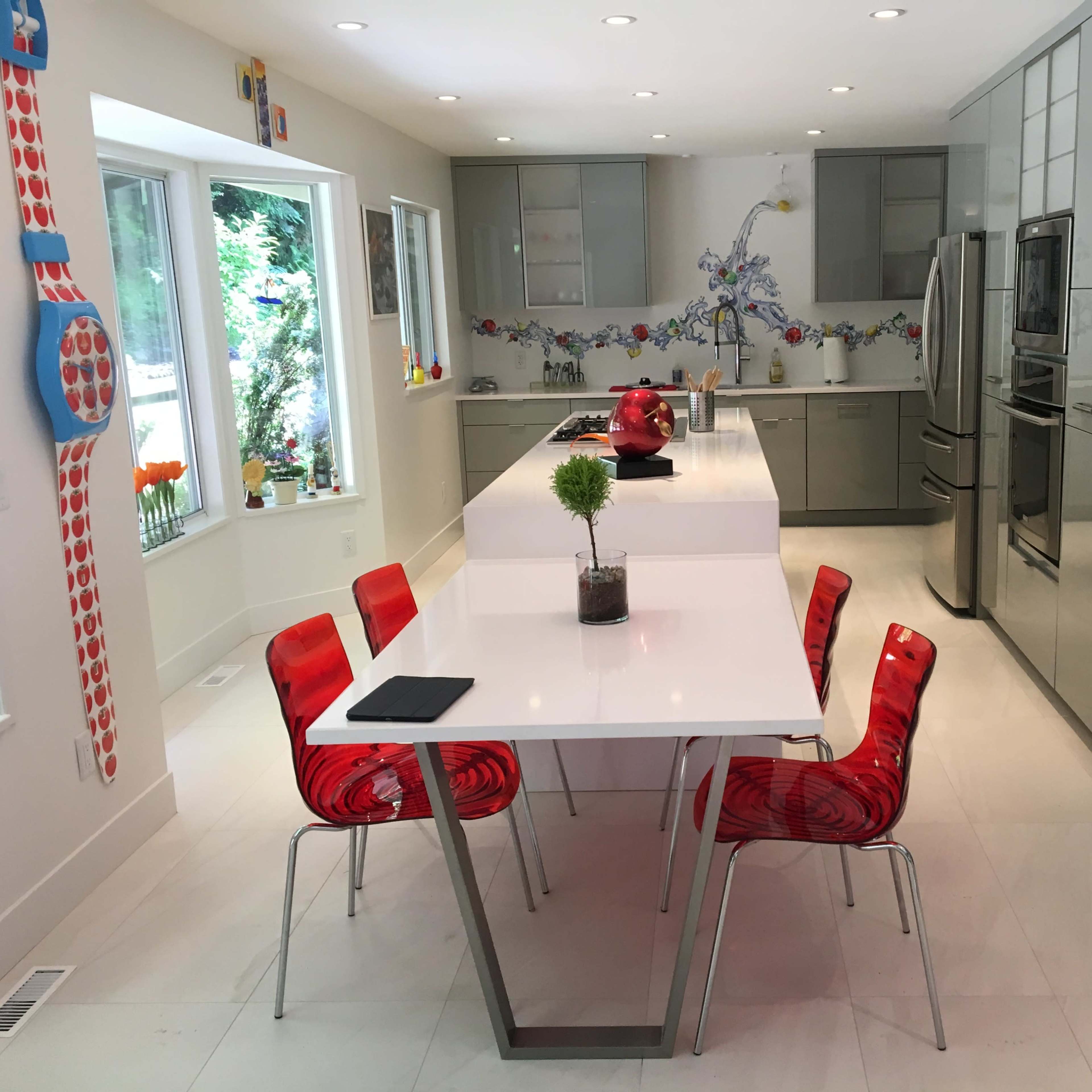 A modern kitchen featuring a sleek white table surrounded by red chairs, with gray cabinetry and stainless steel appliances.