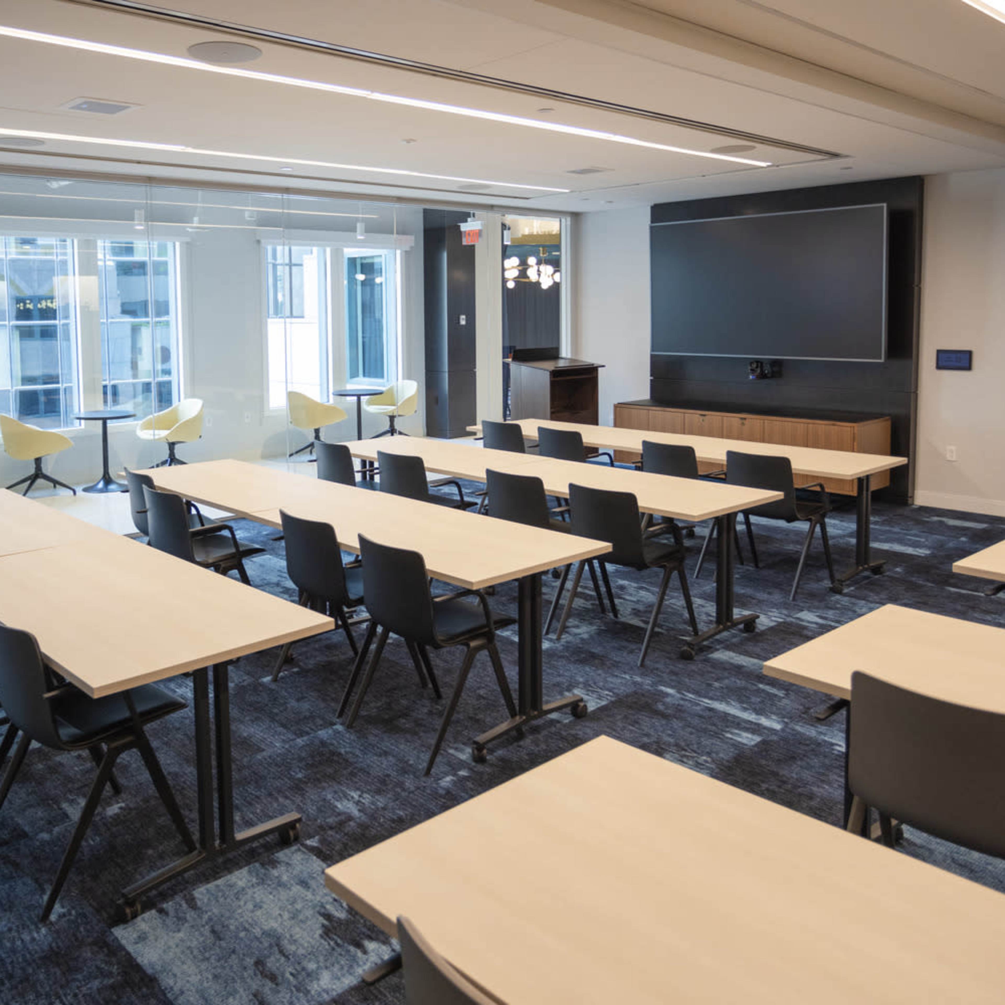 A modern classroom setup with rows of tables and chairs, large windows providing natural light, and a presentation screen at the front.
