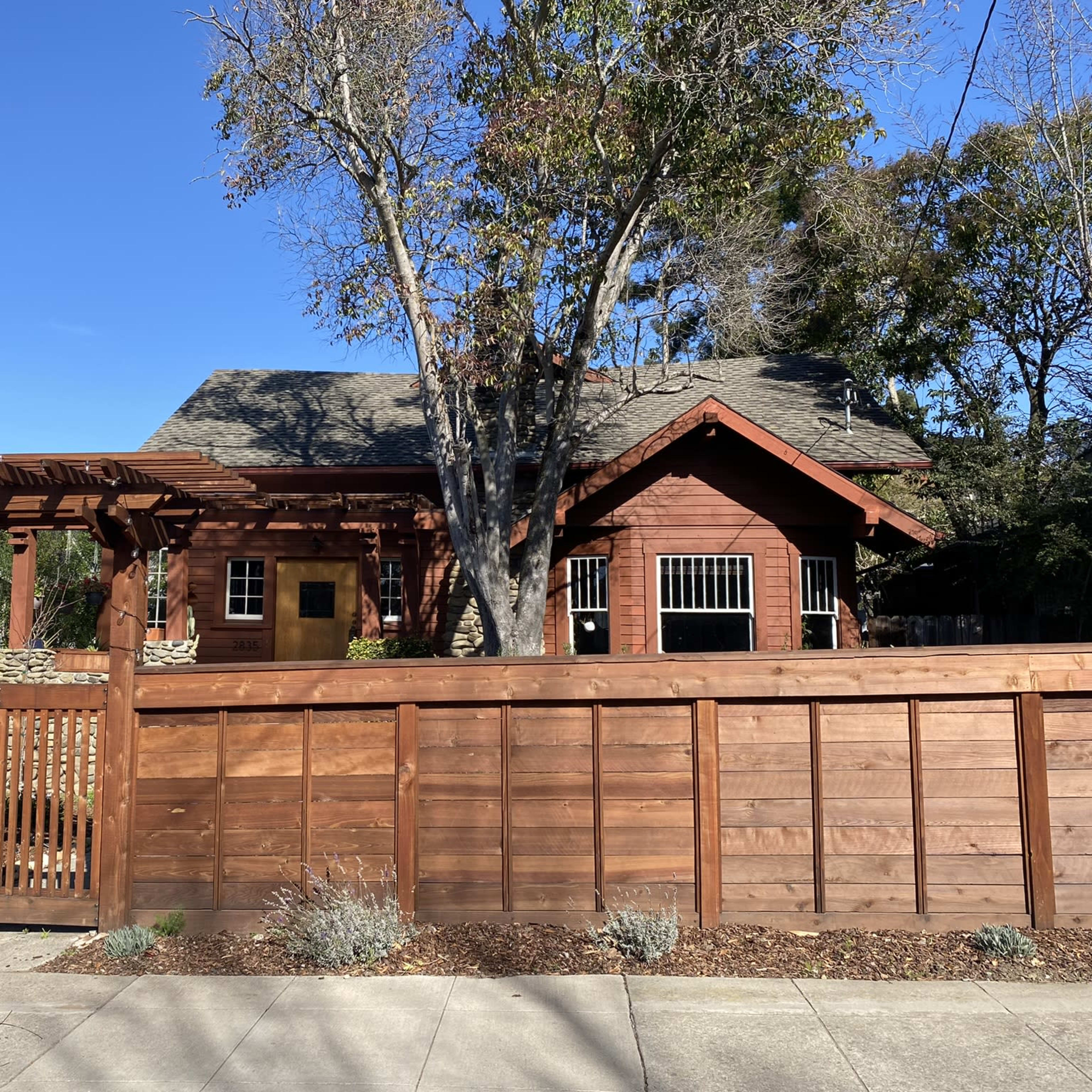 A wooden house with a pitched roof is surrounded by a low fence and features a pergola at the entrance.