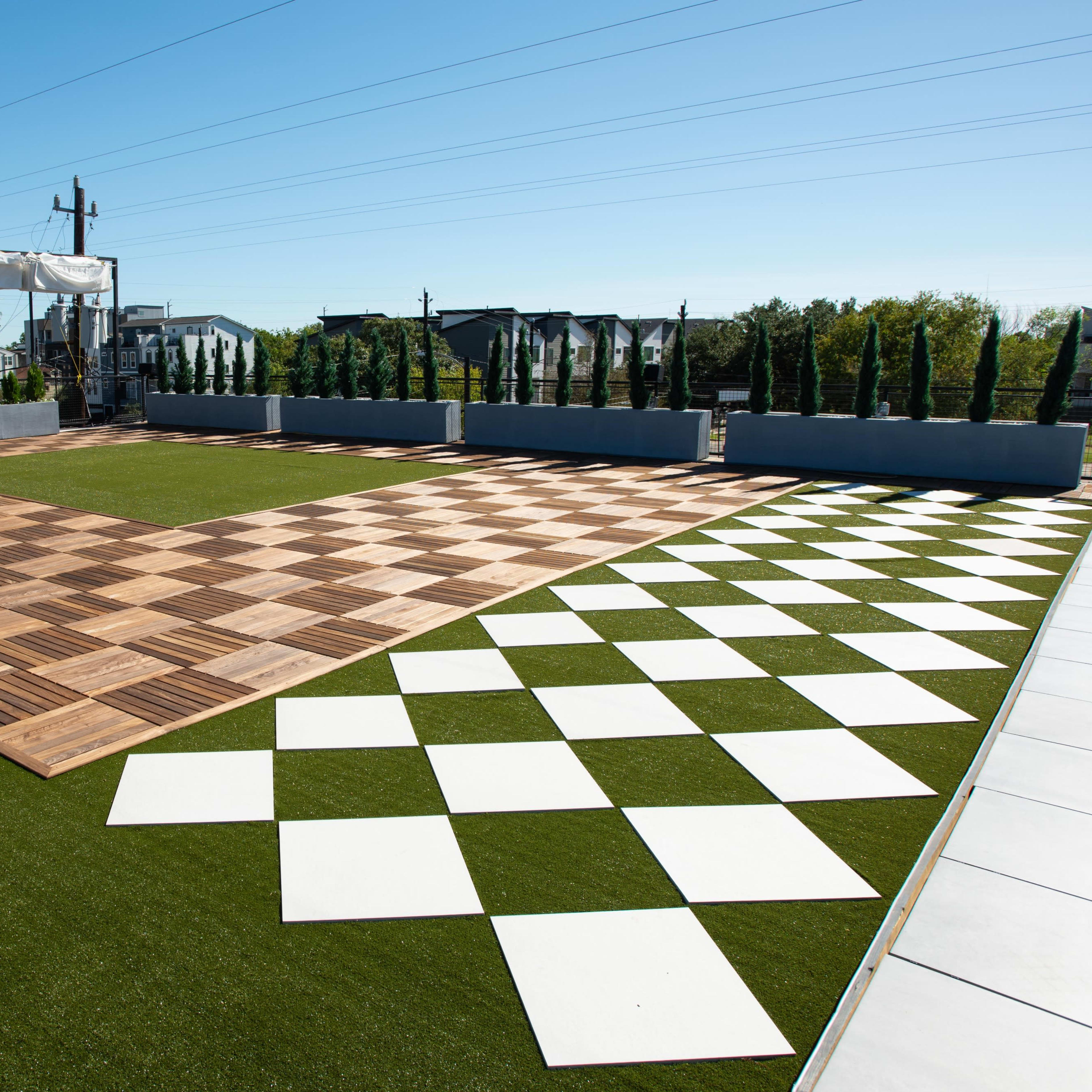 A large outdoor space with a checkerboard-patterned flooring made of wooden tiles and white panels, surrounded by green grass and decorative trees.